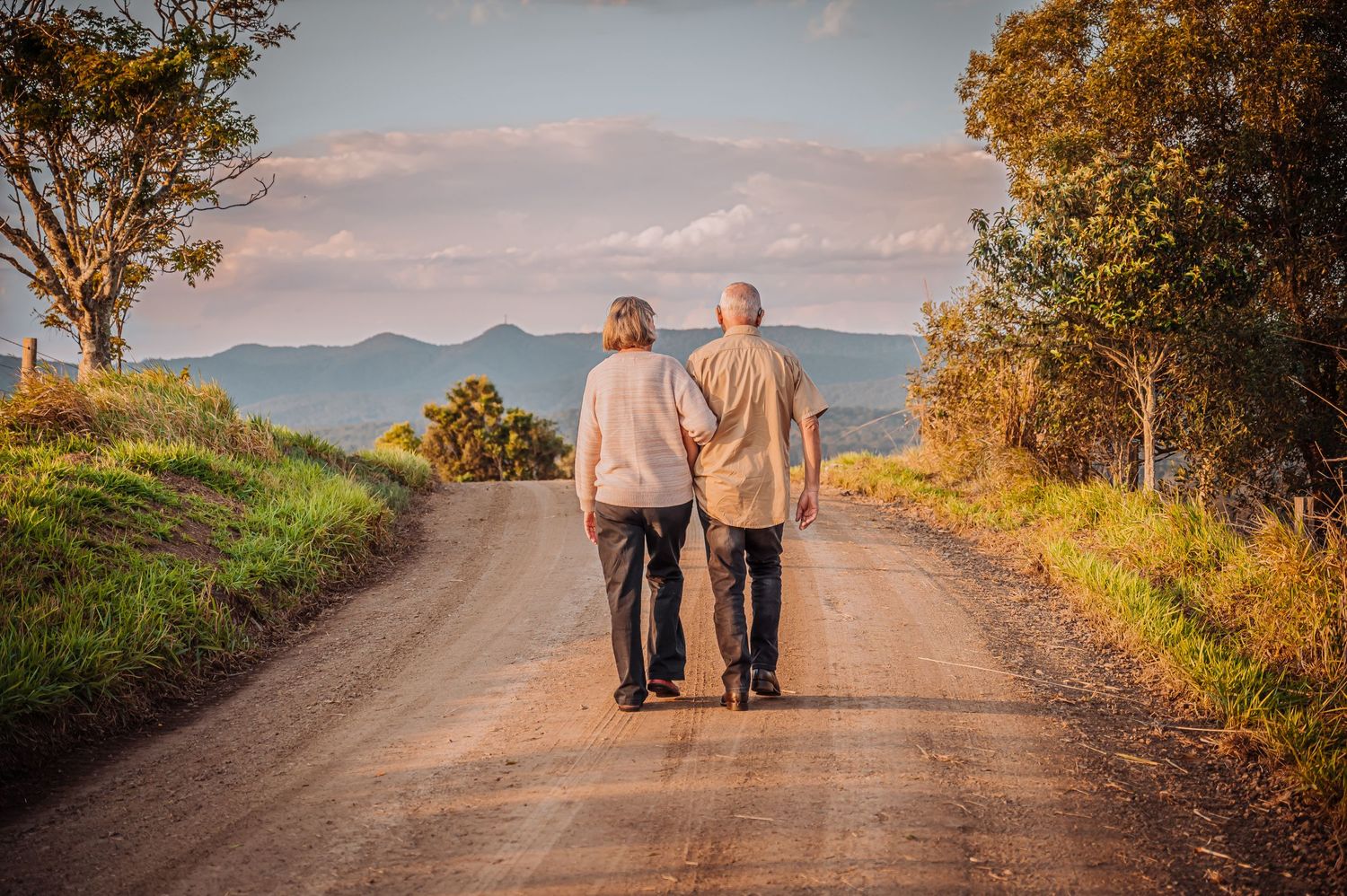 A couple in casual attire walks together down a countryside dirt road at sunset with mountains in the background.