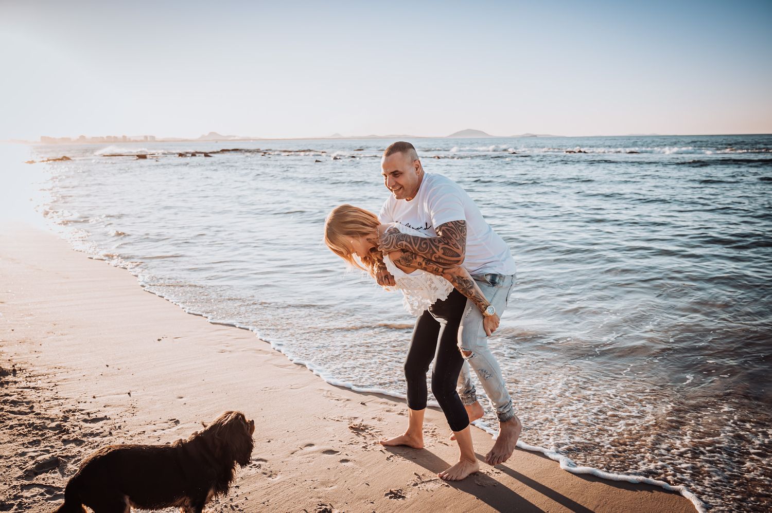 A playful couple enjoys a moment together on the beach with their dog at sunset.