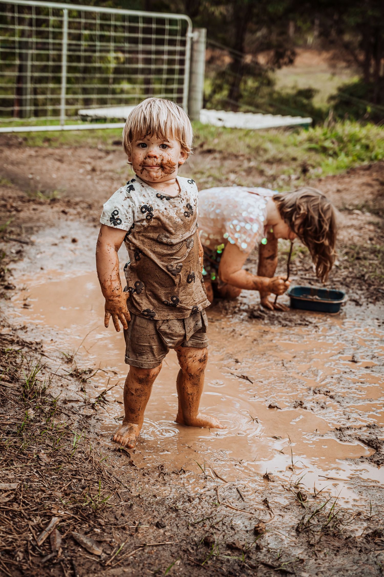 Child playing in a muddy puddle wearing brown overalls on outdoor terrain.