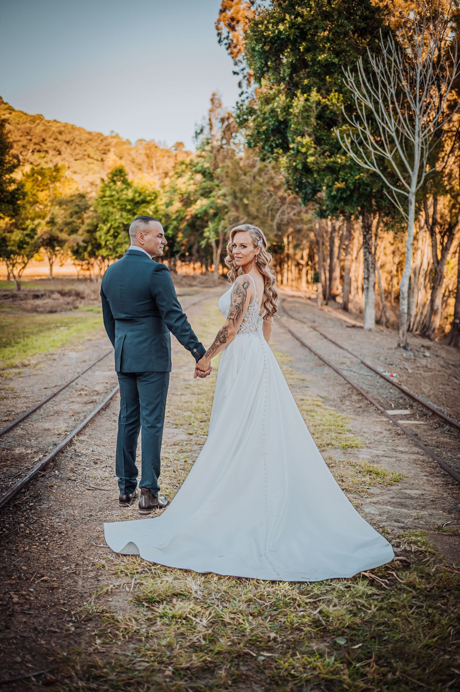A romantic wedding portrait of a couple walking on a tree-lined path with the bride's white dress trailing behind them.