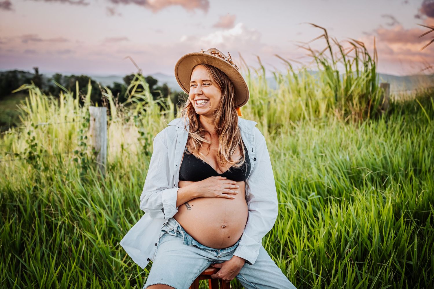 Expectant mother in denim and hat poses in tall grass during golden hour sunset.