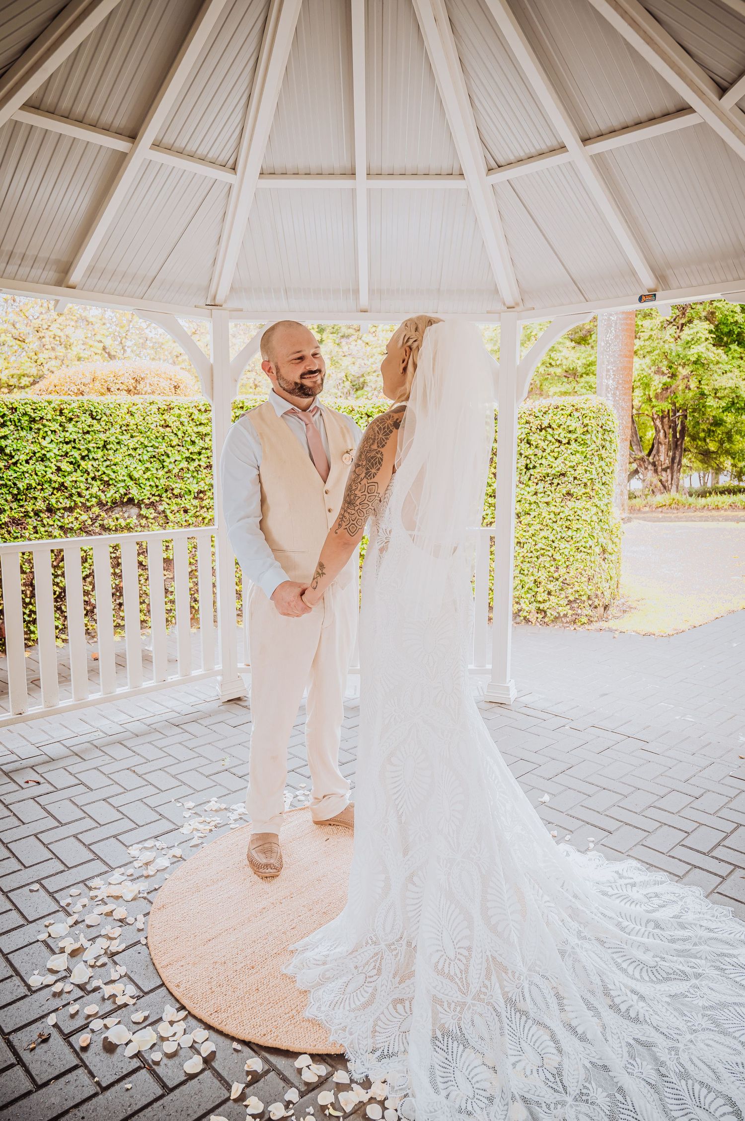 Couple in all-white attire sharing joyful moment in outdoor gazebo with long wedding train.