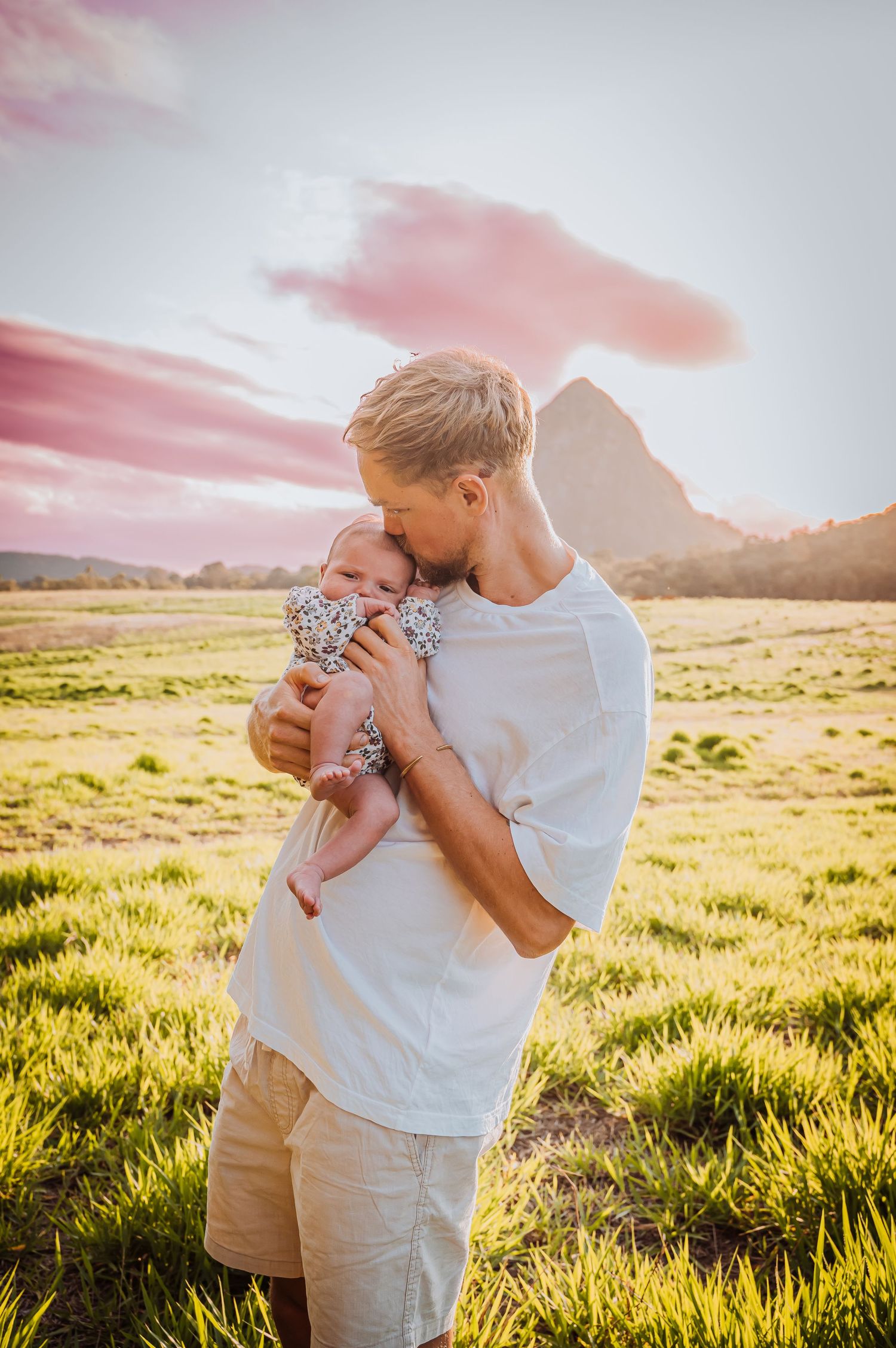 Parent holding infant outdoors in grassy field with mountain and pink sunset sky.