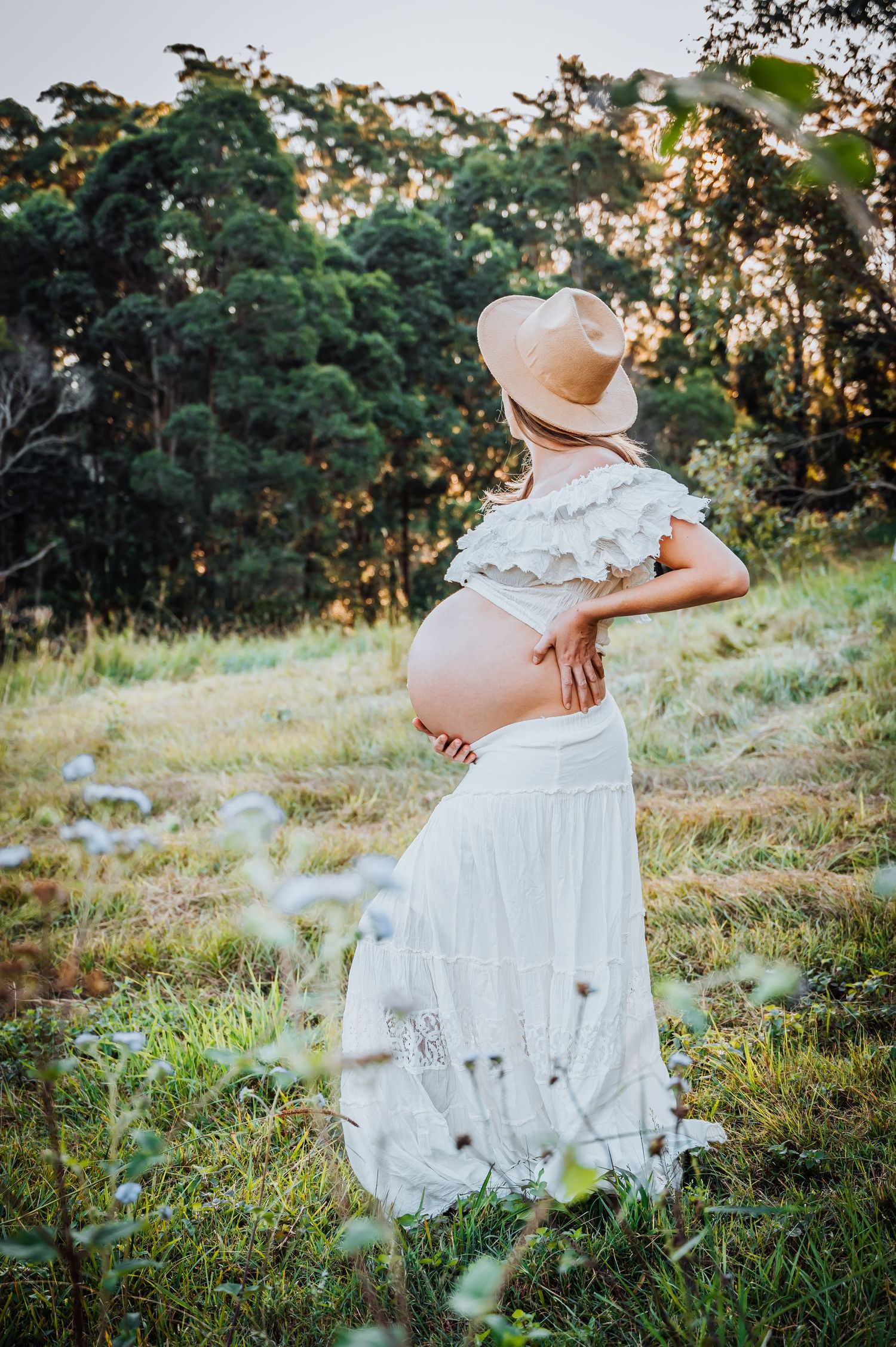 Rustic maternity photo with white dress and straw hat in sunlit meadow at dusk.