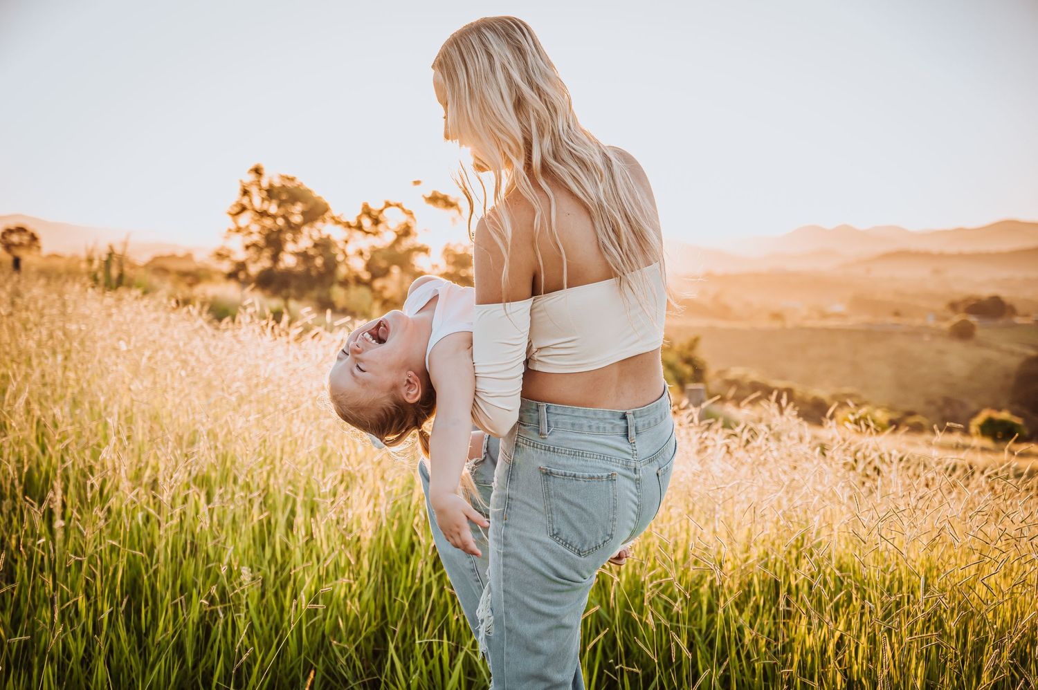 Silhouette against sunset of person holding child upside down in tall grass meadow.