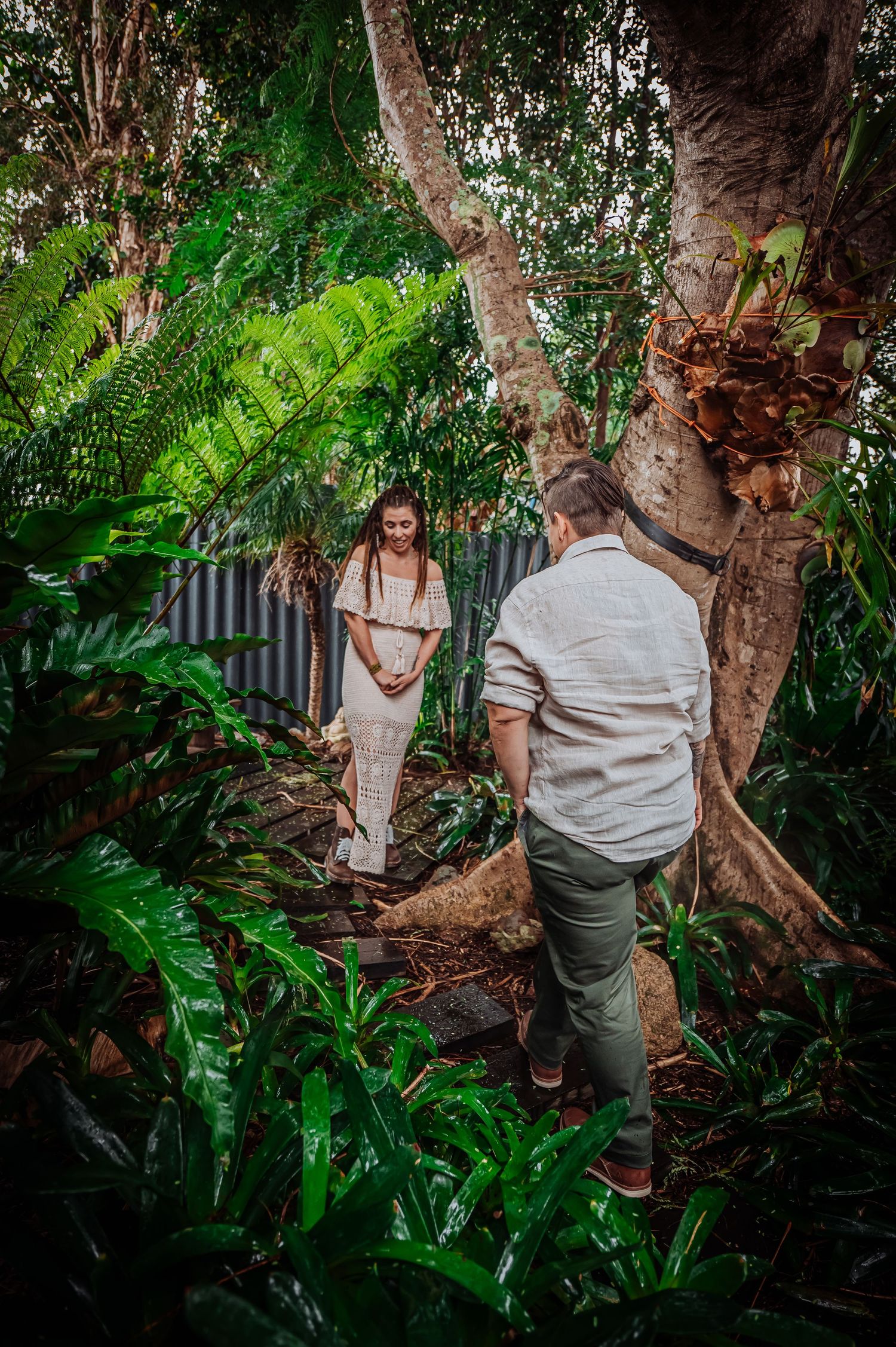 A couple shares a romantic moment among lush tropical greenery and tree trunks.