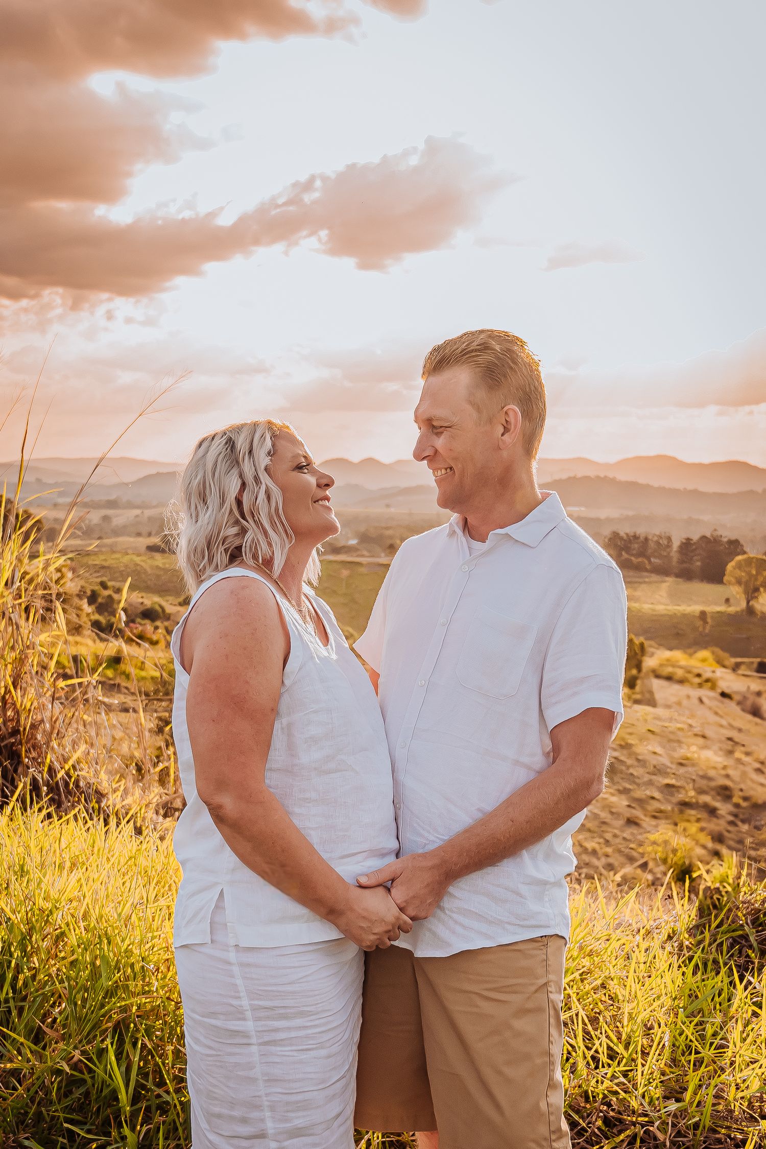 A couple in white outfits shares an intimate moment against a sunset landscape with rolling hills.