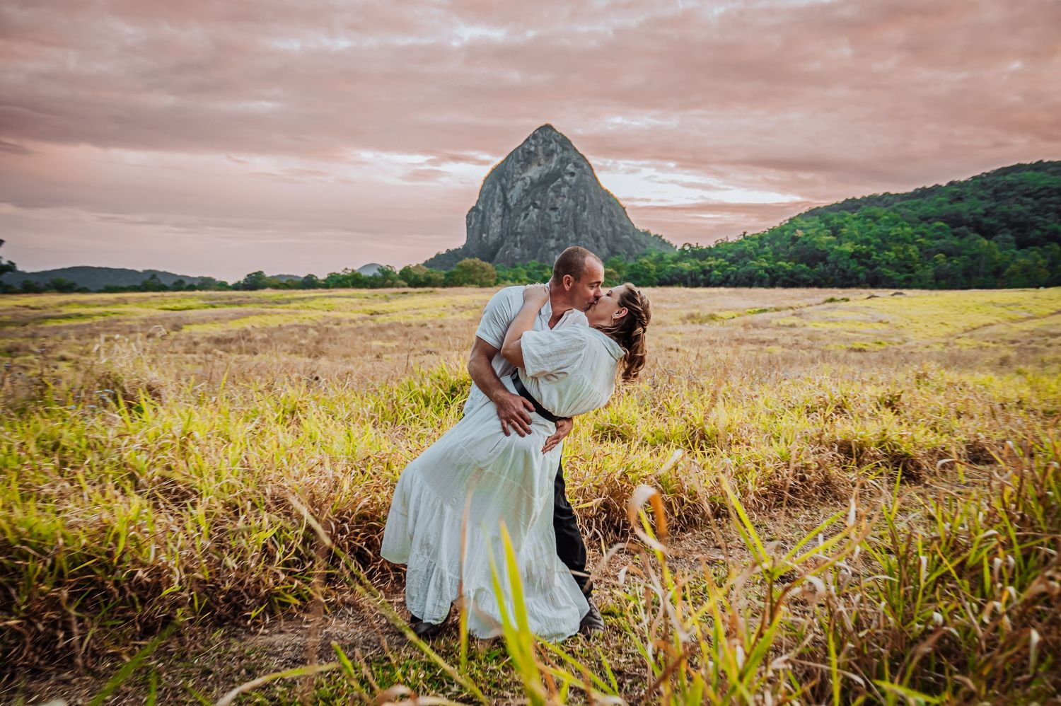 A romantic couple poses dramatically in a golden field with a mountain backdrop during sunset.