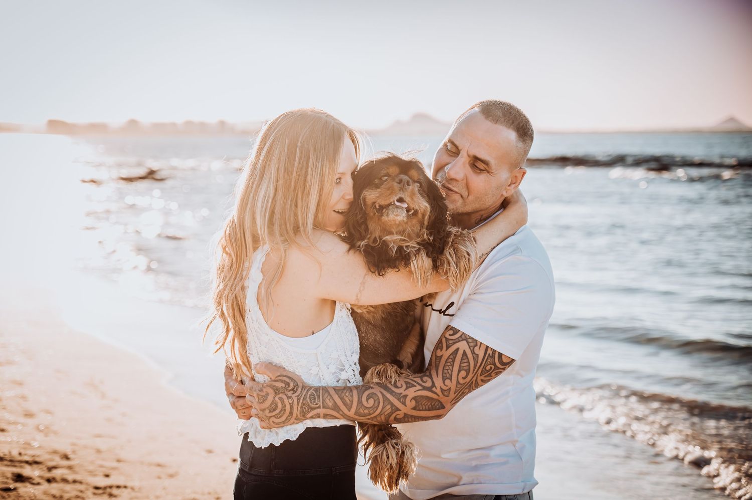 A couple holds their chocolate brown dog together at the beach during sunset.