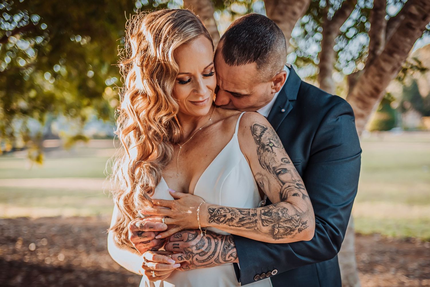 Newlyweds embrace under tree with bride showing intricate sleeve tattoos in white dress.