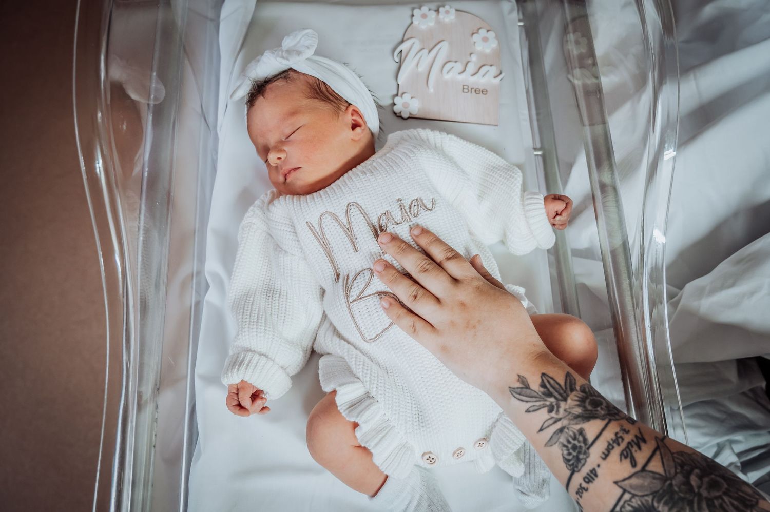 Newborn baby sleeping in hospital bassinet wearing white knit outfit.
