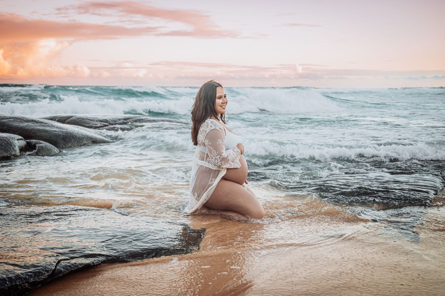 A maternity photo on a rocky beach at sunset with waves crashing against the shore.