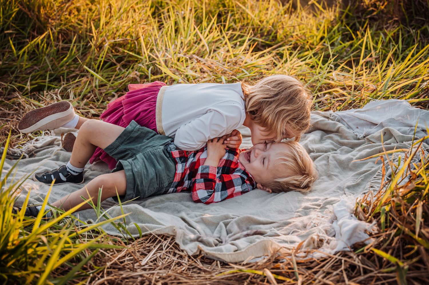Two children in casual wear share a tender moment lying on a blanket surrounded by golden grass at sunset.