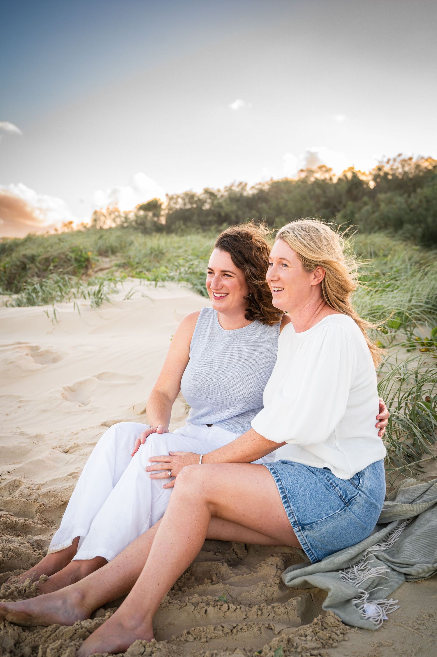 Two friends share a laugh while sitting together on coastal sand dunes at sunset.