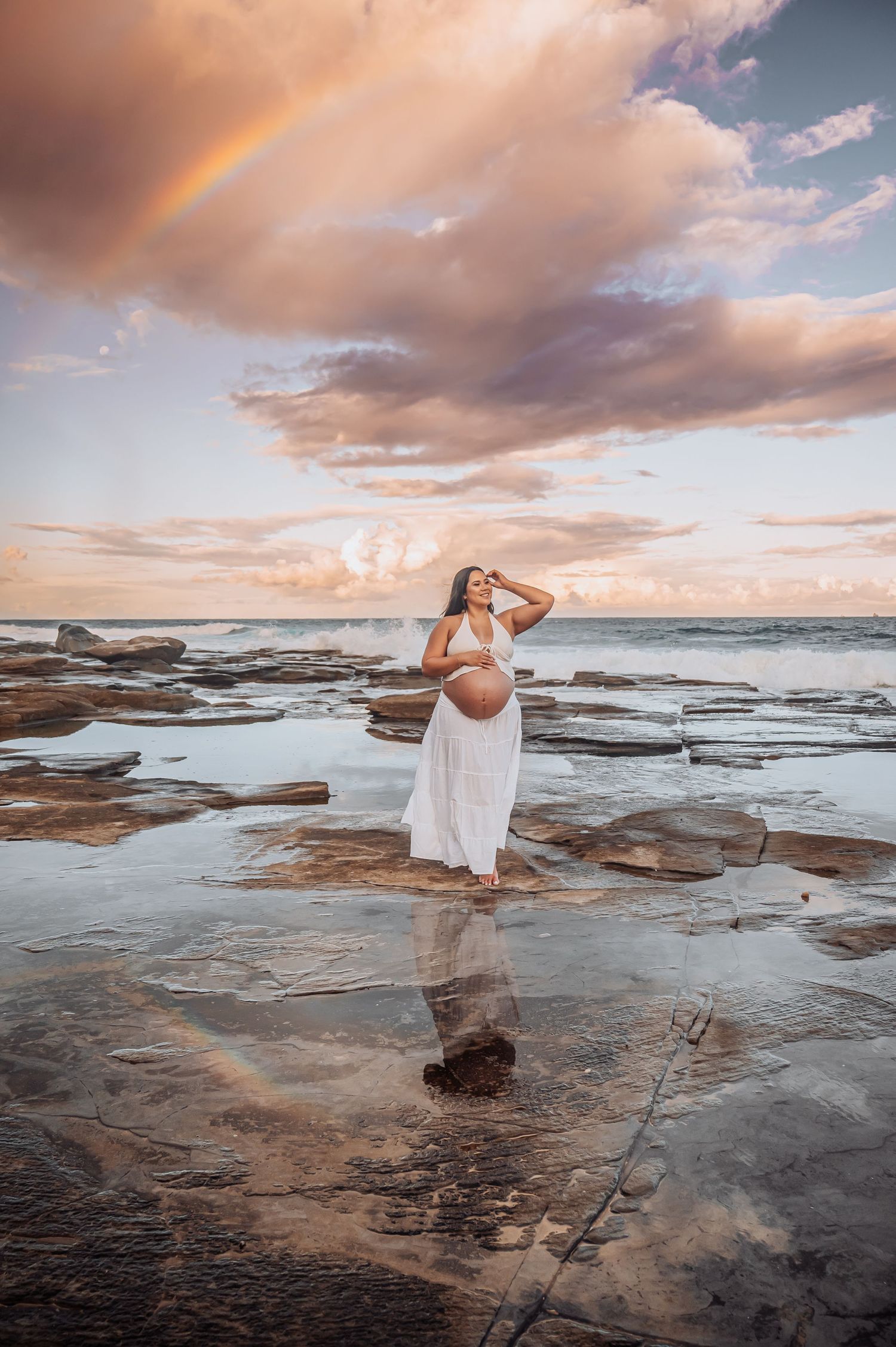 Maternity silhouette against dramatic sunset sky with rainbow over rocky coastal landscape.
