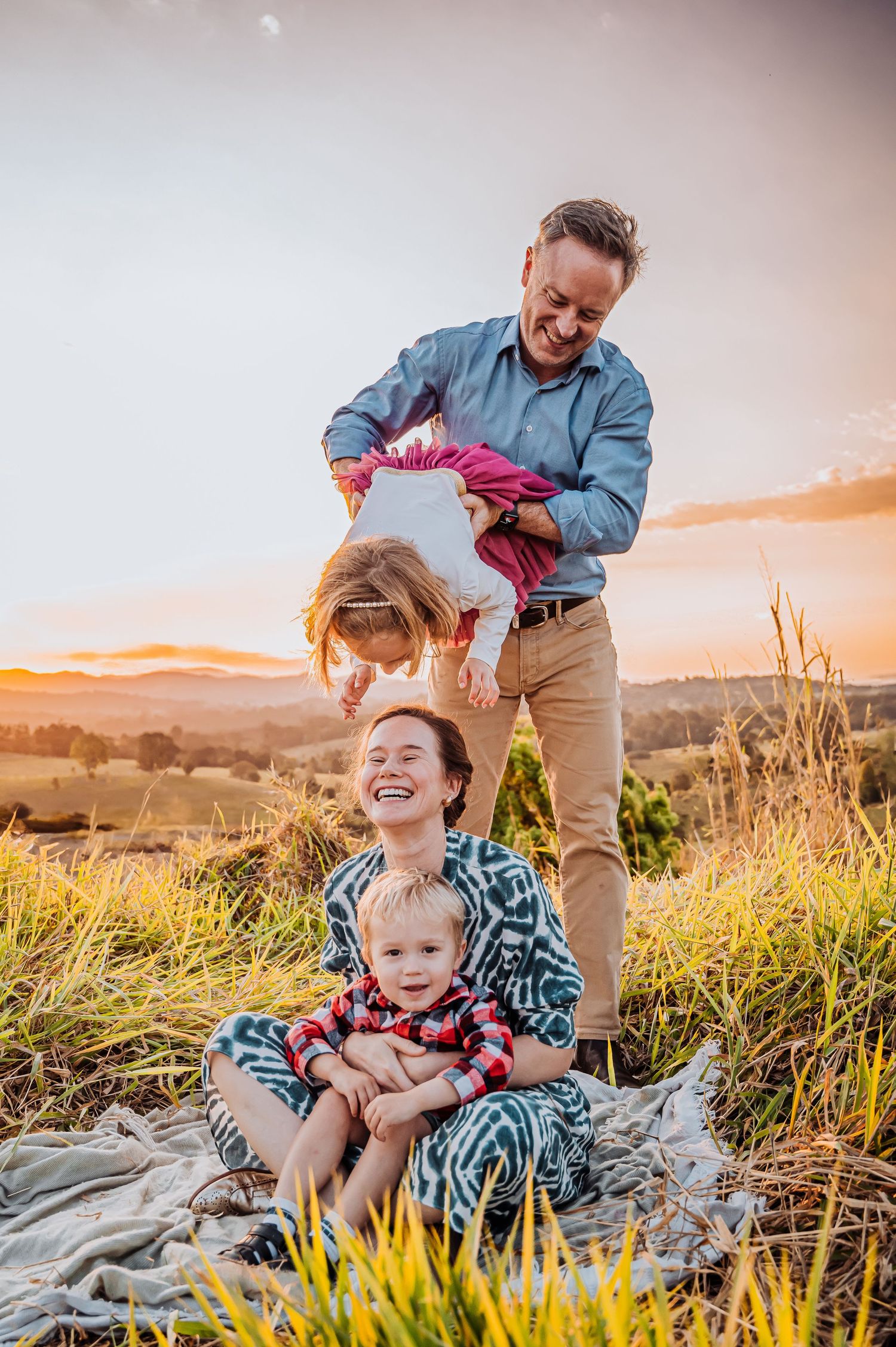 Family enjoying playful moments together on grassy hillside during golden hour sunset.