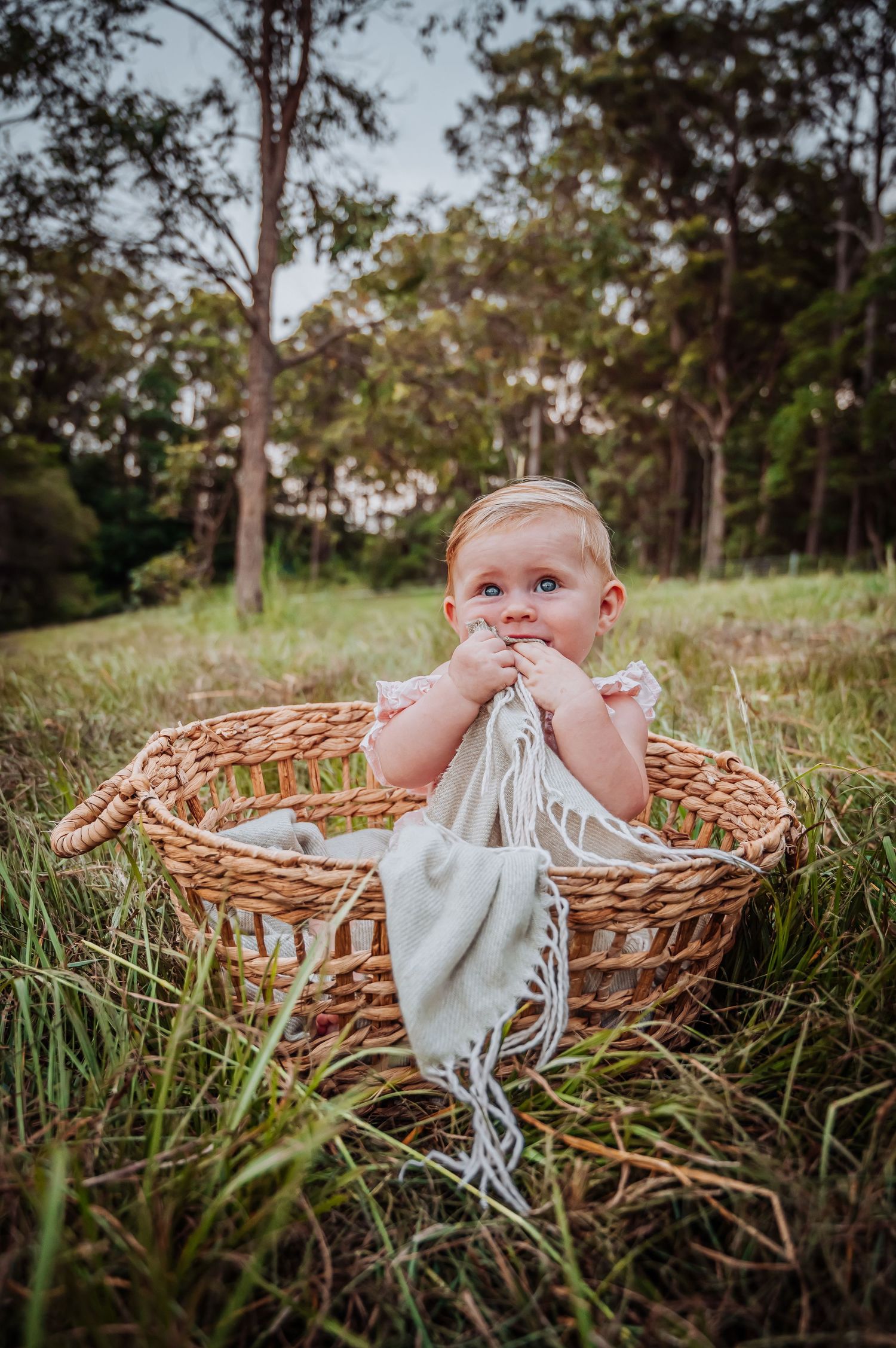 Small child in white outfit sitting in woven basket while playing with fabric in grassy field.