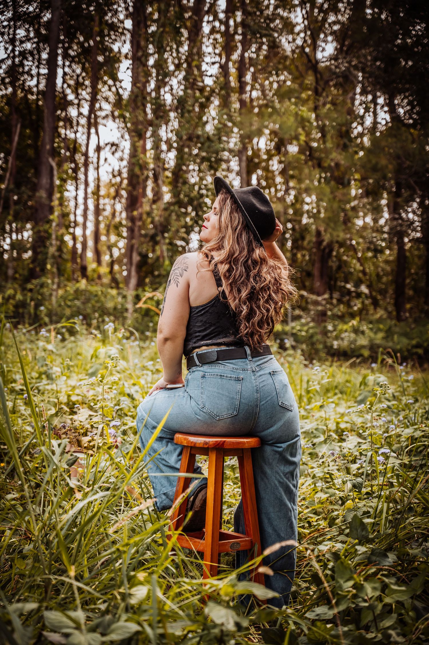 Natural light portrait of person in hat and jeans seated on wooden stool surrounded by forest foliage.