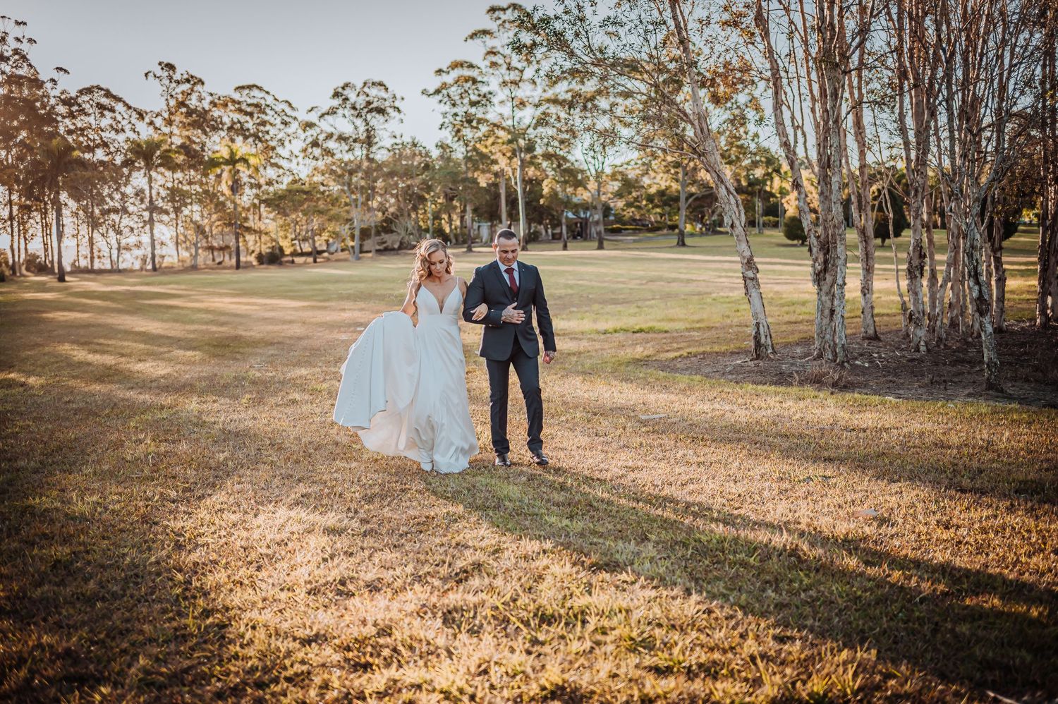 Bride in flowing white gown walks with groom across sunlit field with eucalyptus trees.