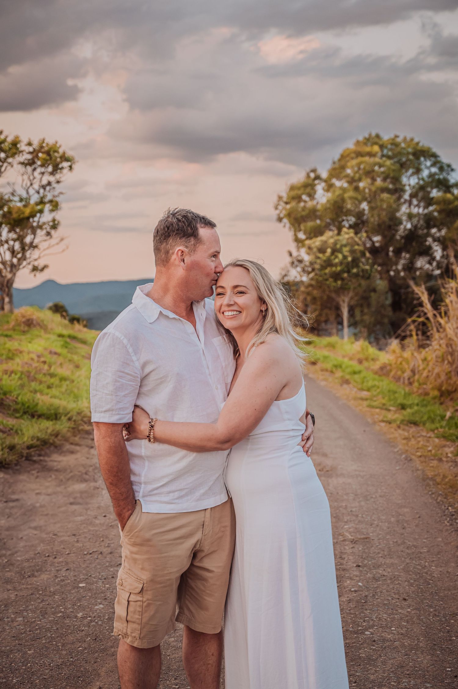 A romantic couple embraces on a dirt road at sunset with moody clouds overhead.