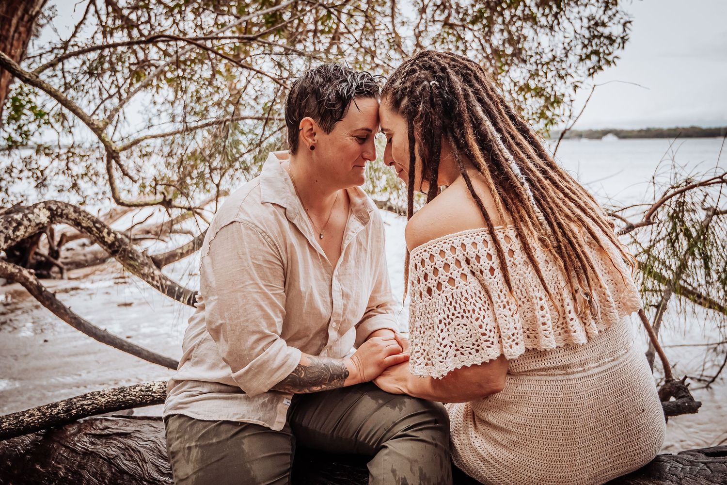 Intimate moment between couple sitting on driftwood by the water in matching beige outfits.