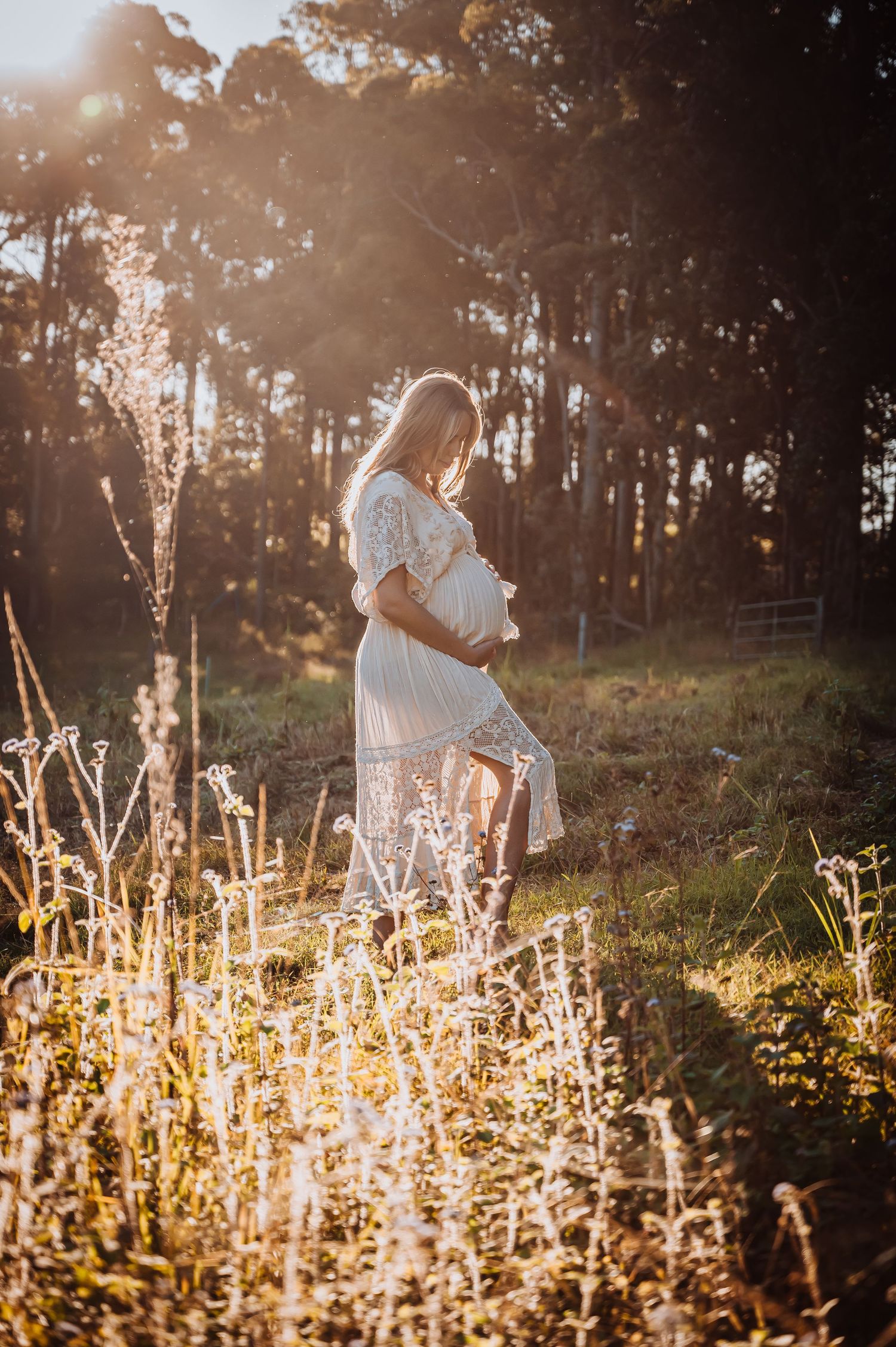 Ethereal maternity photo of an expectant mother in a white lace dress standing in a sunlit forest at golden hour.