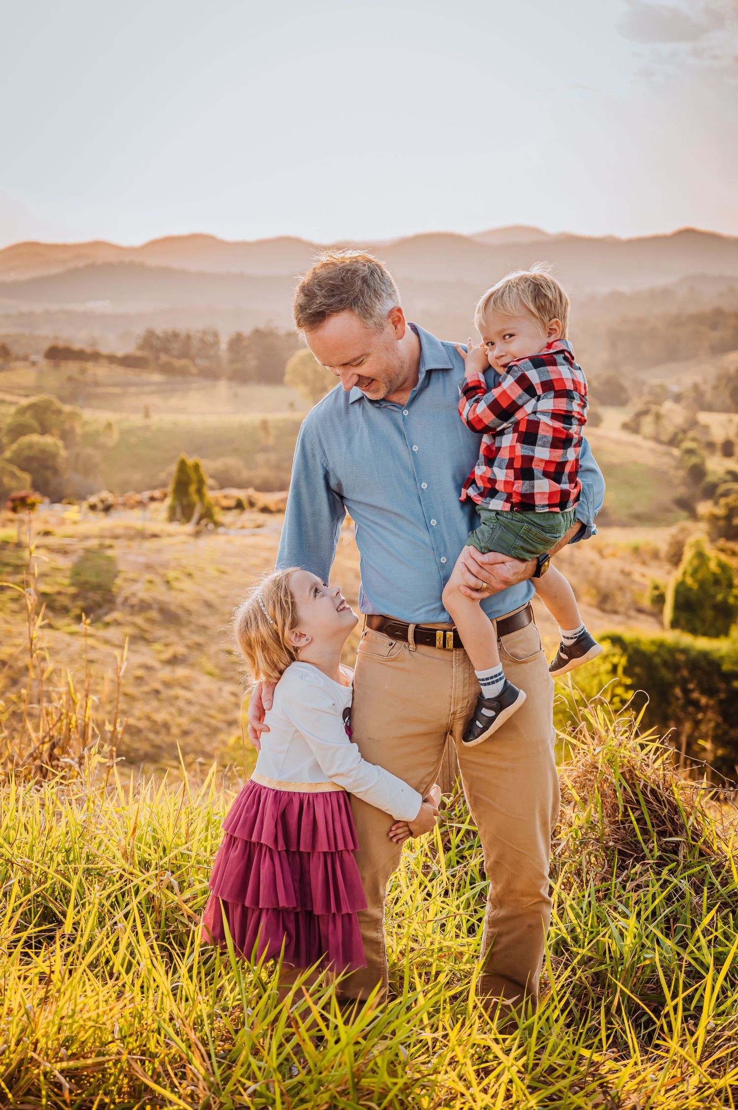 Family enjoying tender moment together in grassy field with mountains in background at sunset.