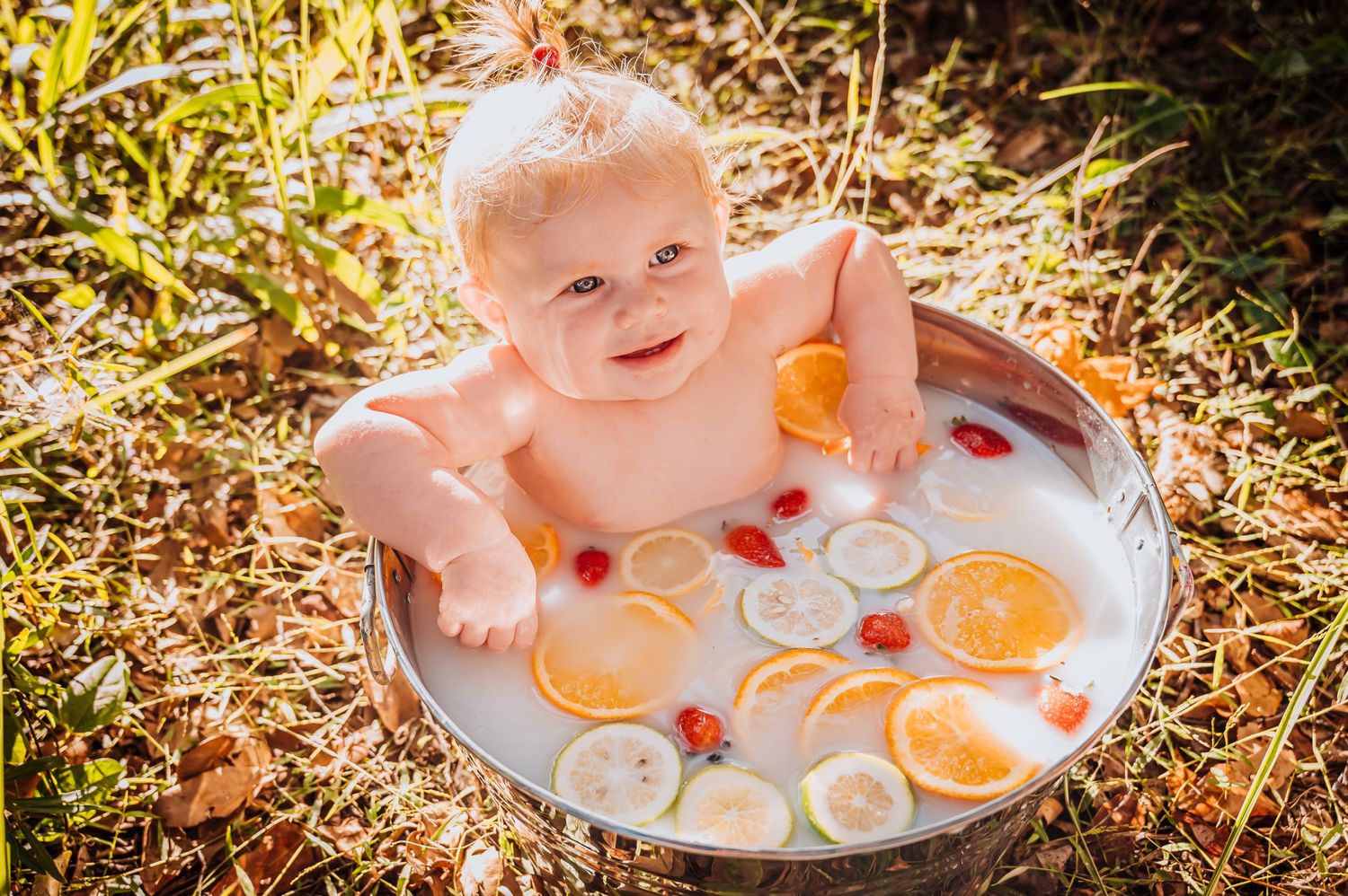 Baby relaxing in citrus milk bath with orange slices and flowers floating in sunlit setting.