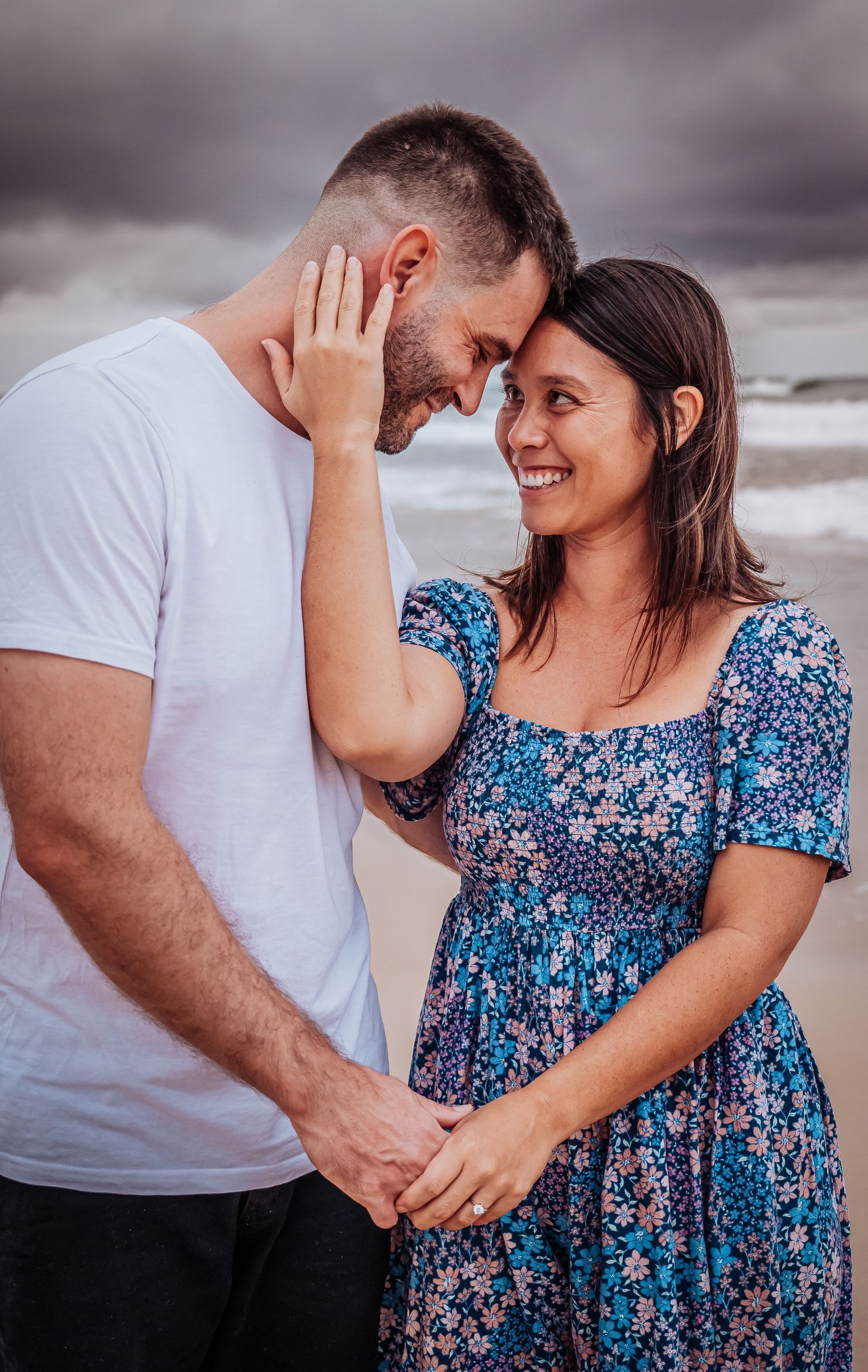 A tender moment shared between a couple at the beach with moody storm clouds in the background.