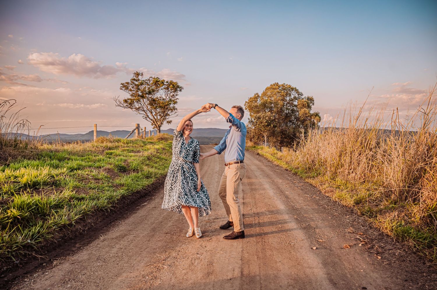A couple dances playfully on a dirt path surrounded by grass and trees at sunset.