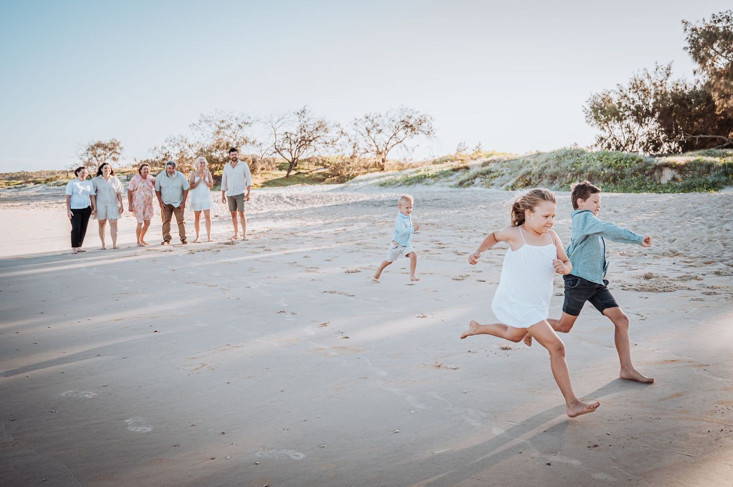 Family members play and run together on a sandy beach at sunset with trees in the background.