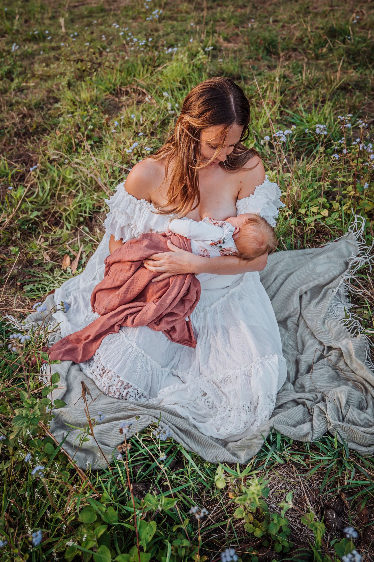 Mother in white lace dress breastfeeding infant on blanket outdoors among wildflowers at sunset.