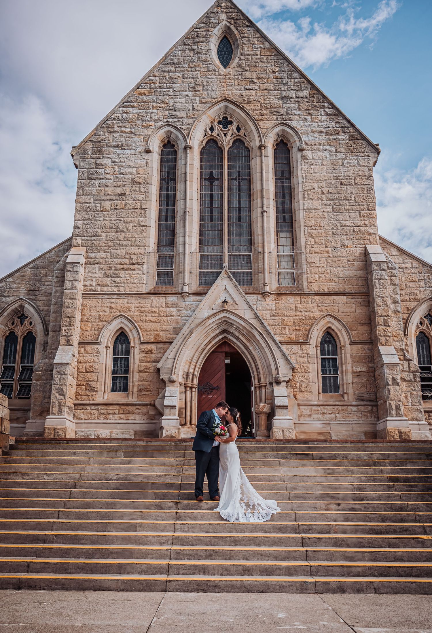 A newly married couple poses on the steps of a grand stone church with Gothic architecture.