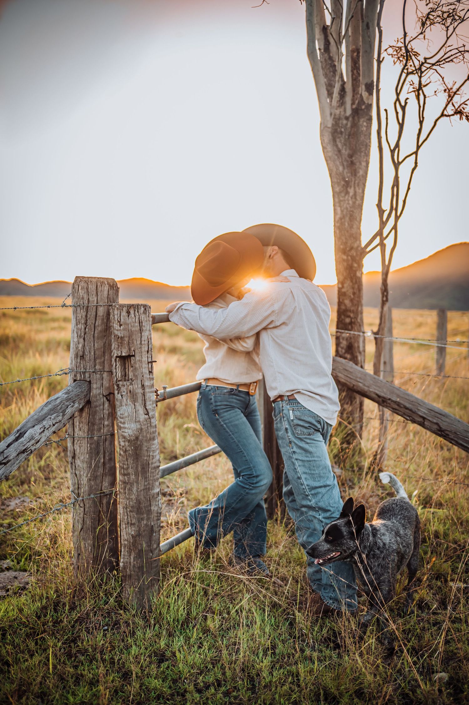 Romantic couple embracing by rustic fence during golden sunset in countryside field.