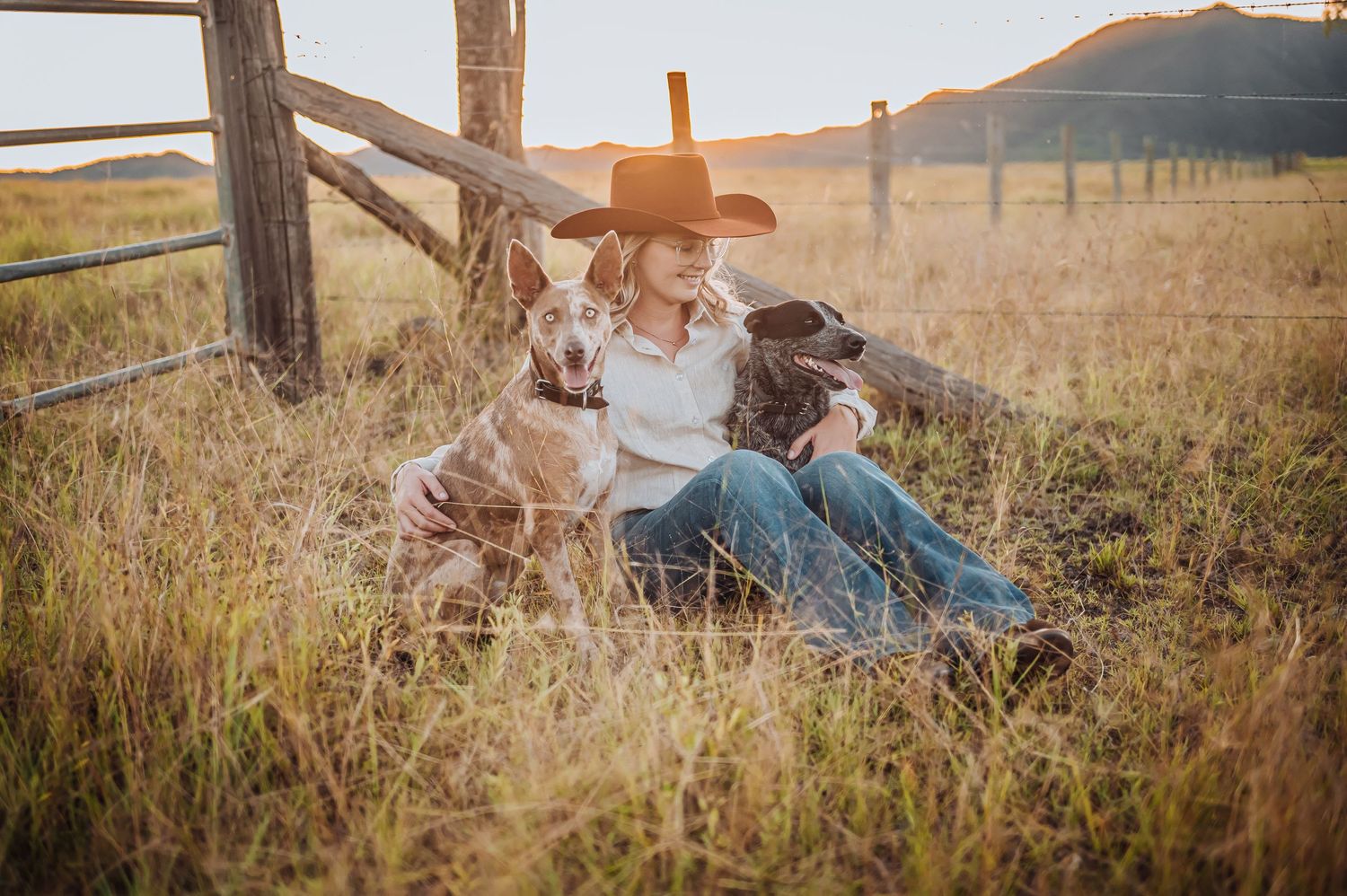 Person in cowboy hat relaxing with two dogs against wooden fence in rustic field setting.