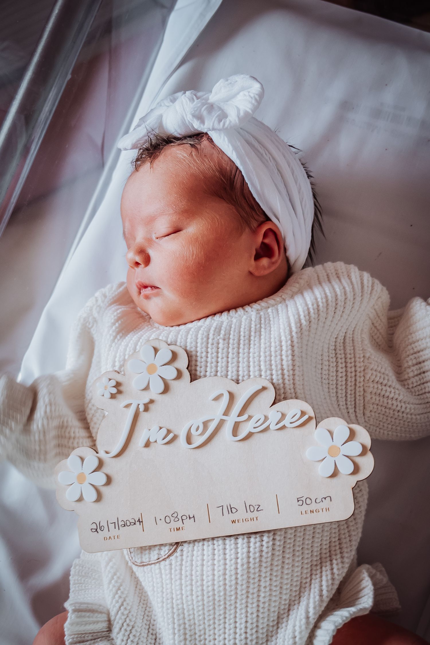A newborn wearing a white knit outfit and headband rests peacefully with a decorative birth announcement.