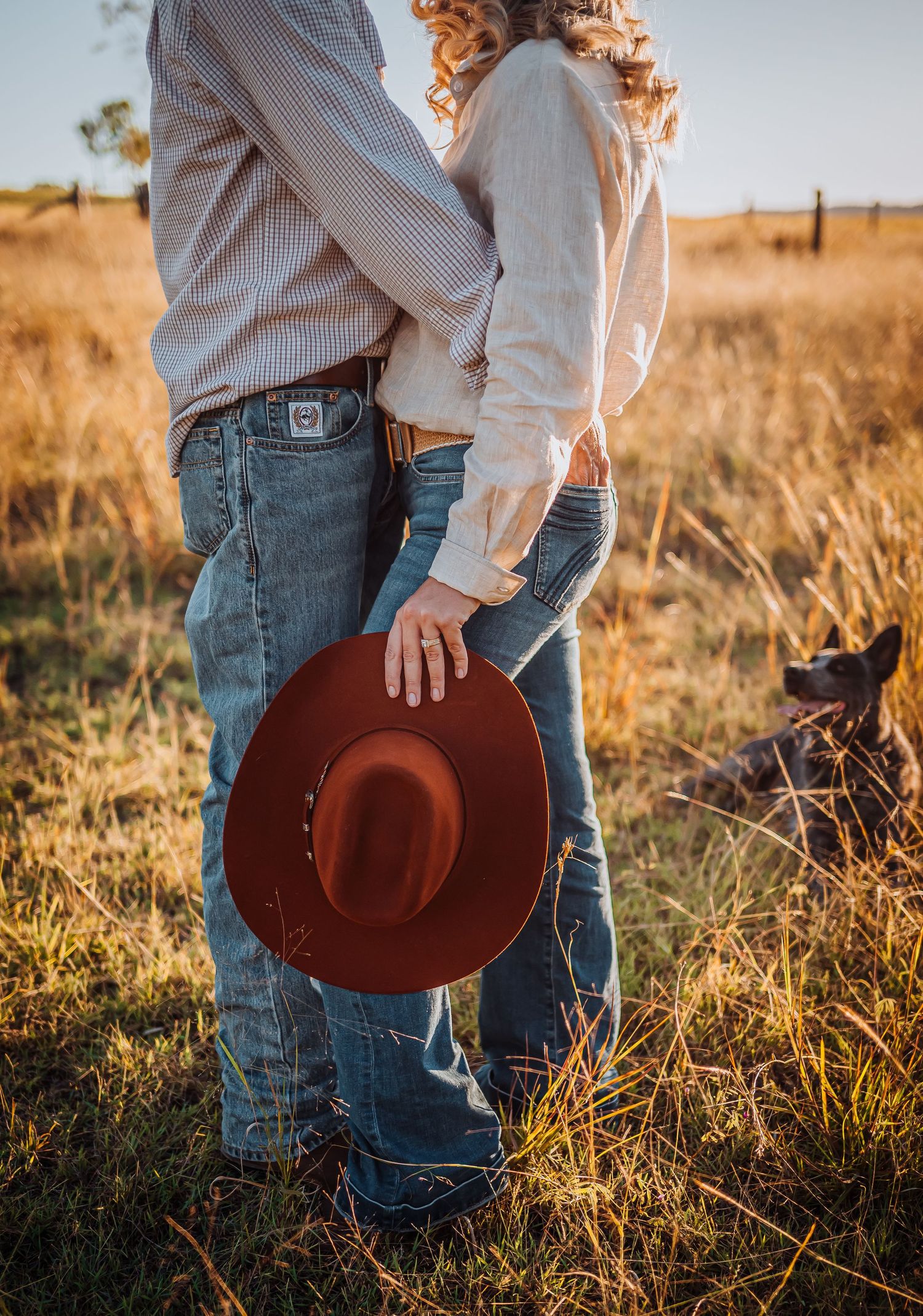 A romantic country scene with denim-clad couple holding a brown felt hat in golden hour light.
