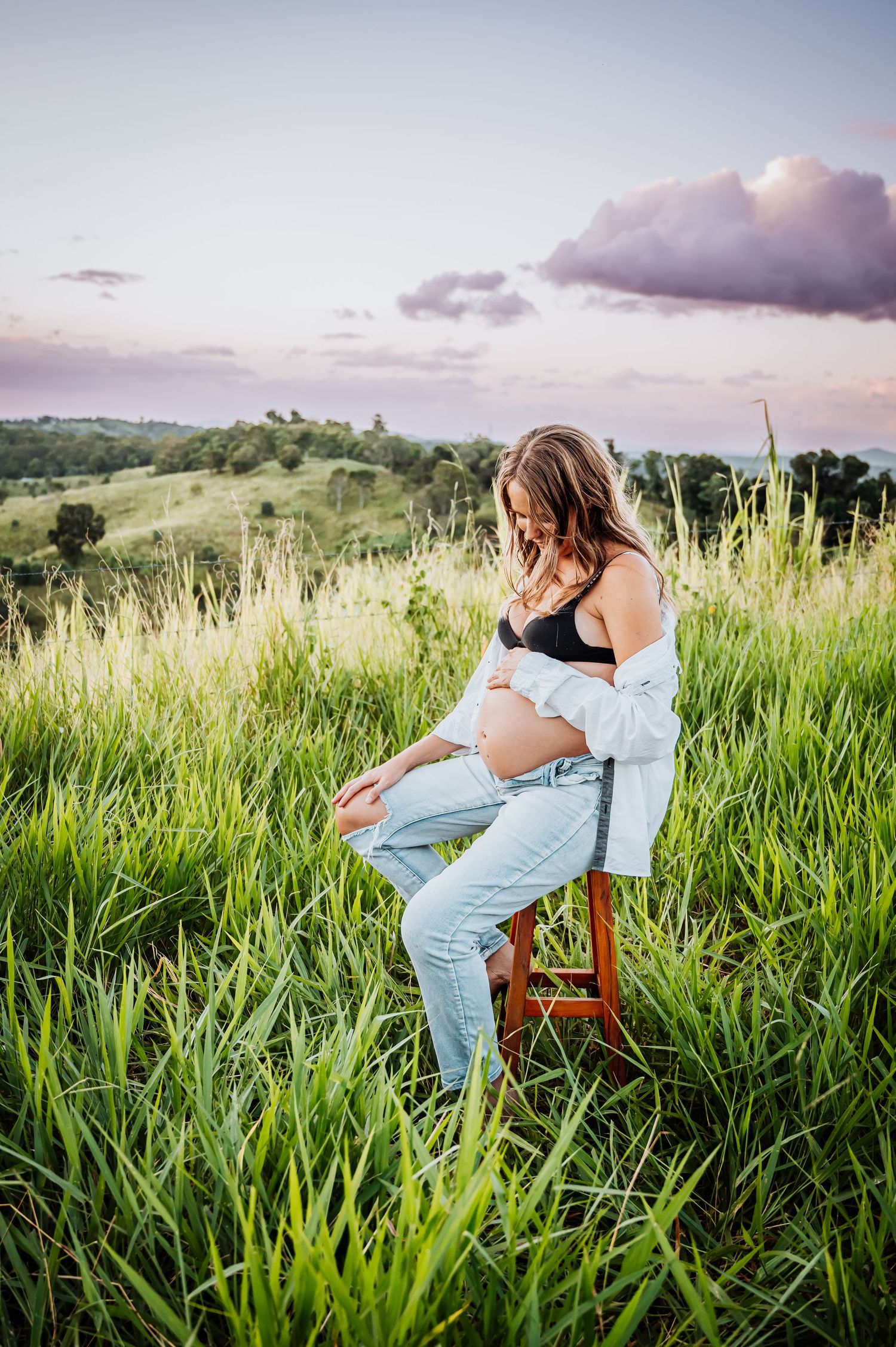 Rustic maternity portrait on wooden stool surrounded by tall grass at sunset.