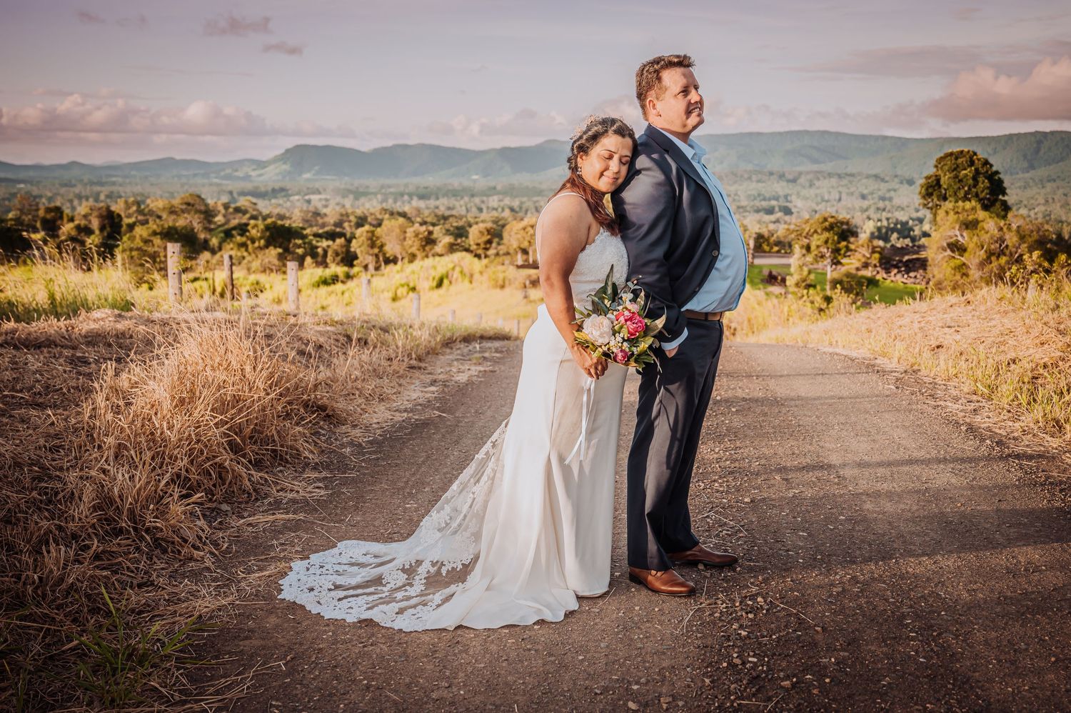 Wedding couple standing on dirt road with mountain vista and sunset sky in background.