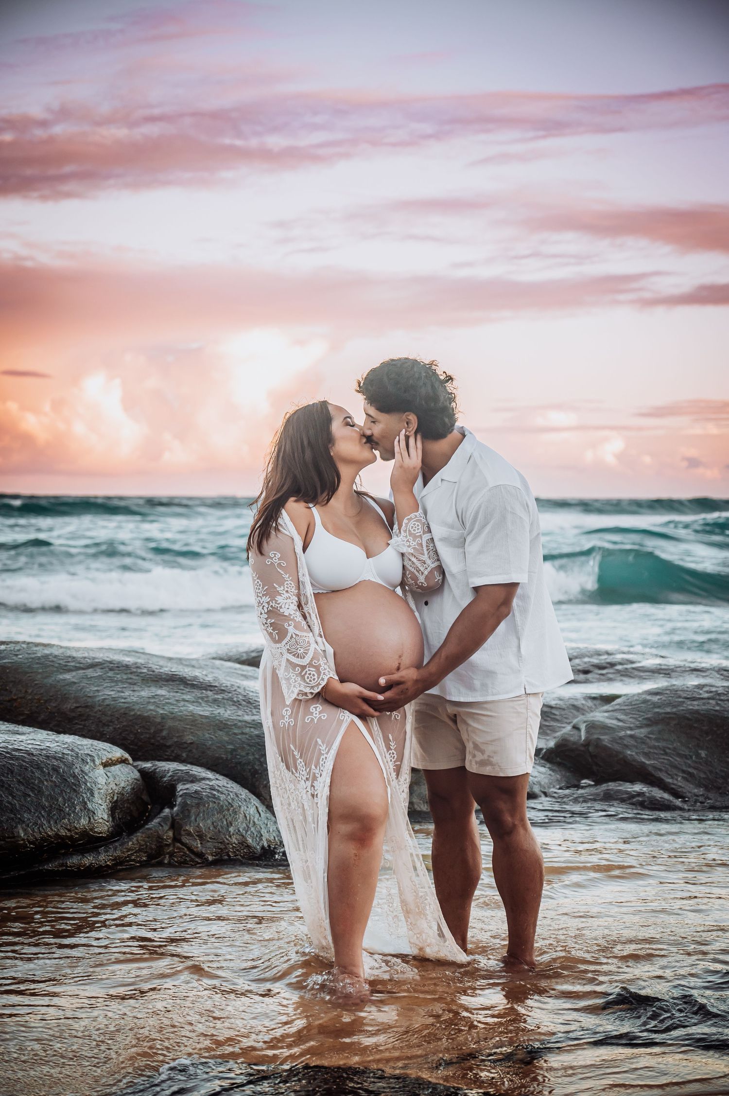 Romantic maternity photoshoot on a rocky beach at sunset with crashing waves.