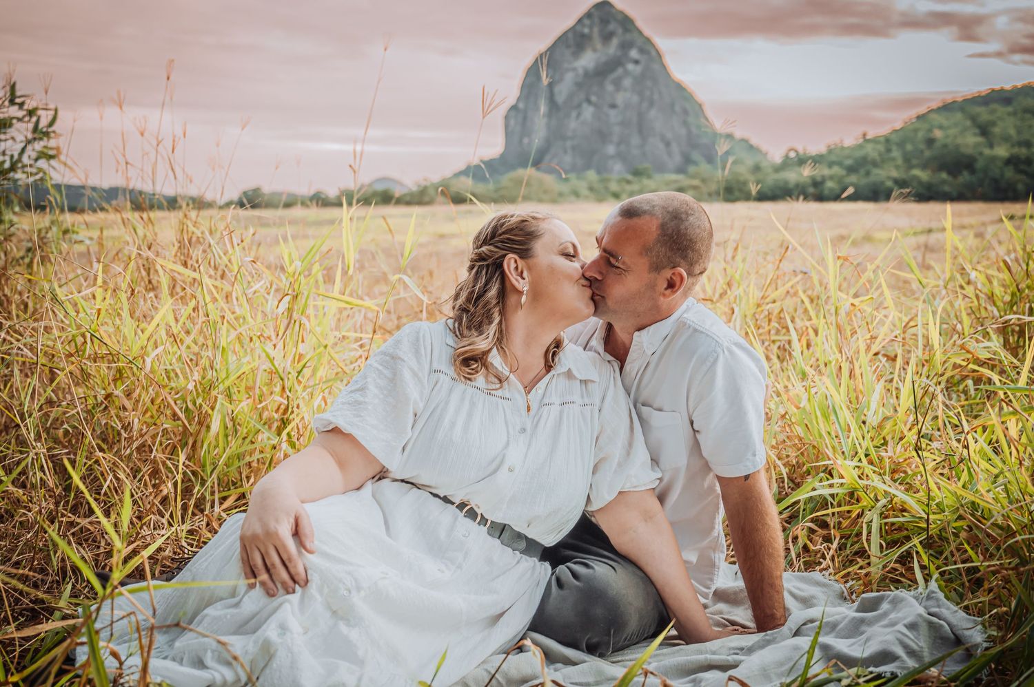 A couple shares an intimate moment while sitting in a grassy field with a distinctive mountain peak in the background.