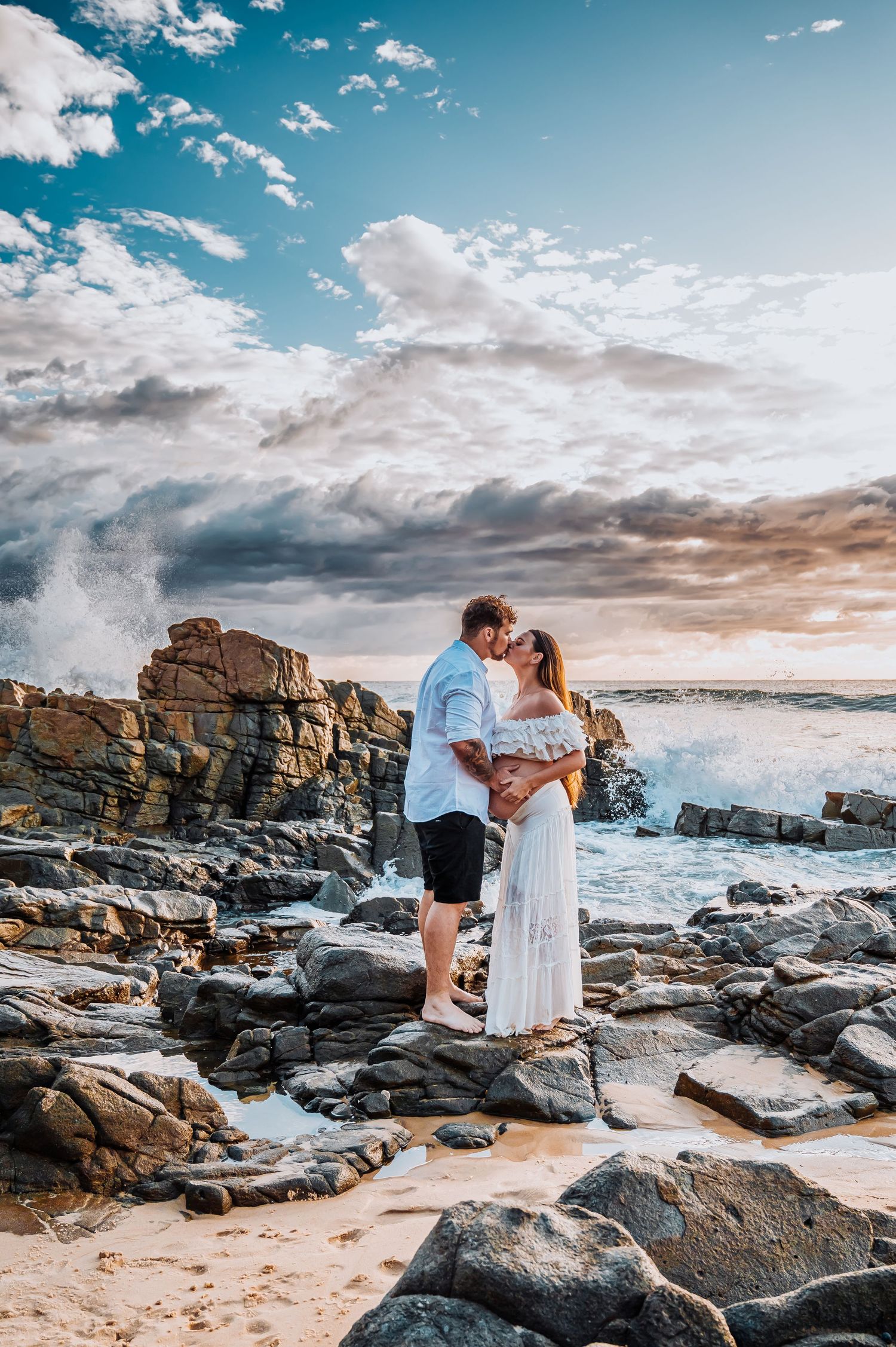 Romantic coastal photo shoot with dramatic clouds and crashing waves on rocky beach.