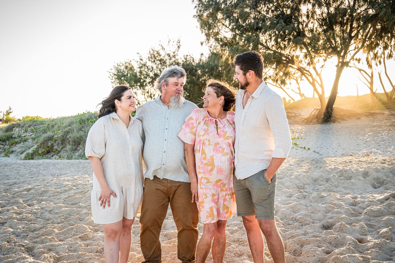 Group shares candid moment together on sandy beach at sunset with trees in background.