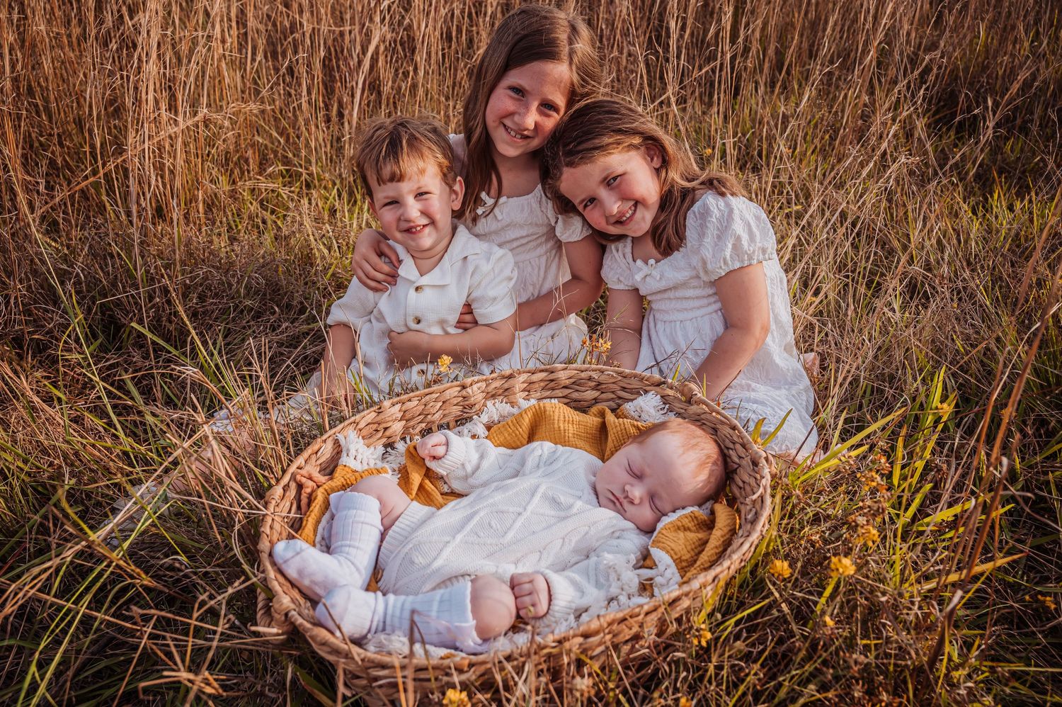 Family gathering around newborn baby in wicker basket outdoors in tall grass at sunset.