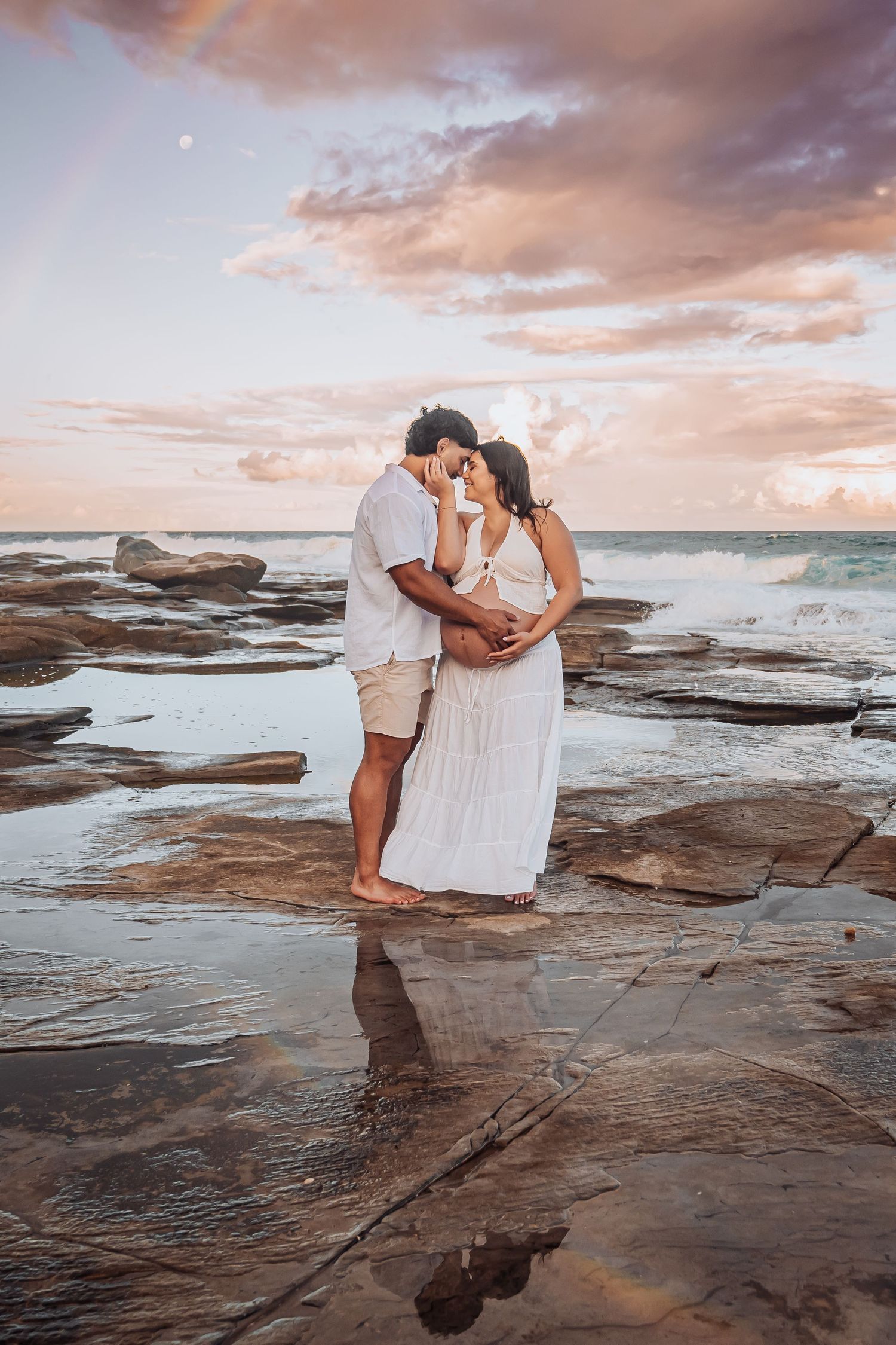 A romantic sunset photo on rocky coastal shores with waves crashing in the background.