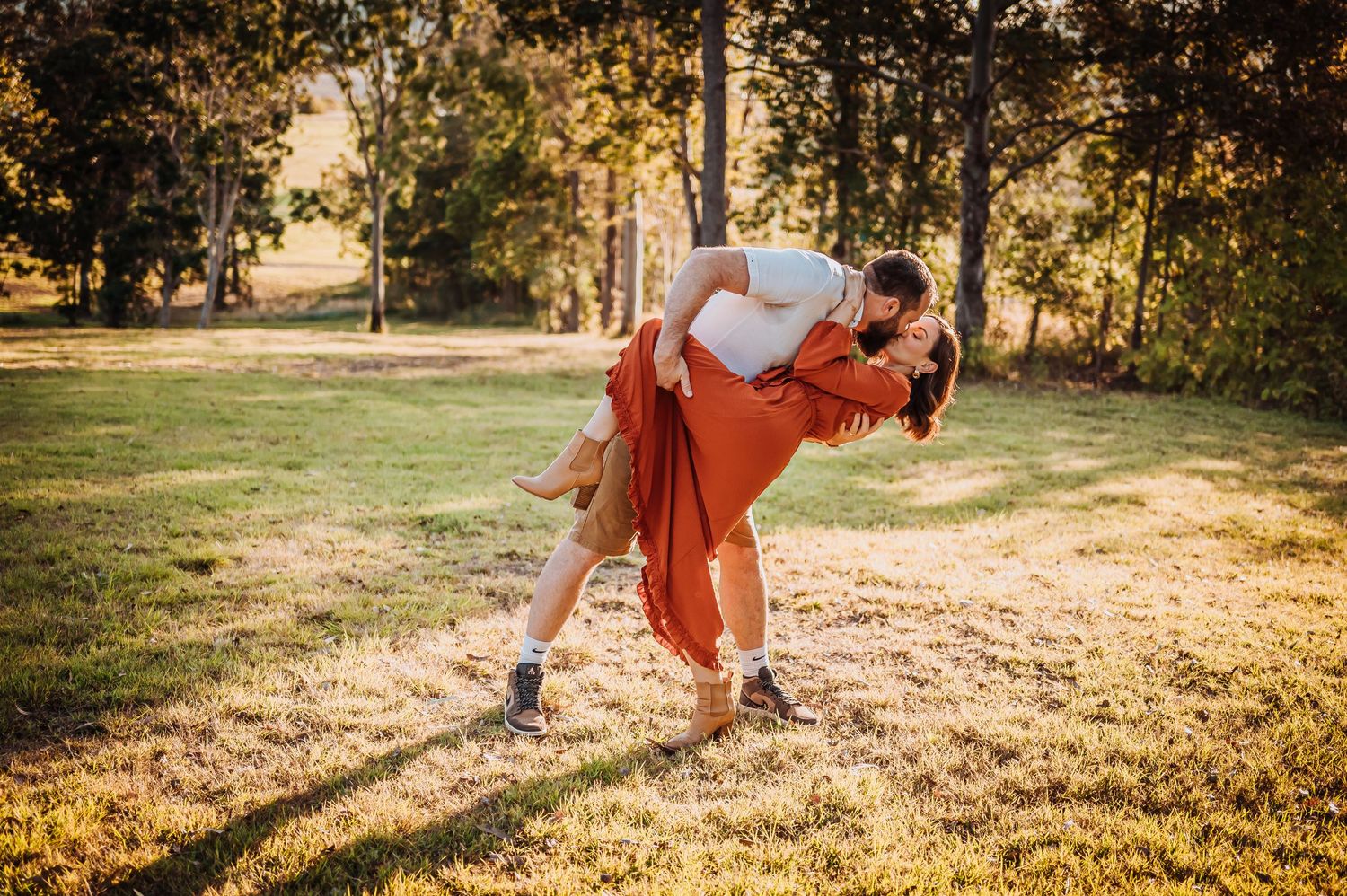 A dancing couple in white and rust colored outfits perform a dip move on sunlit grass.