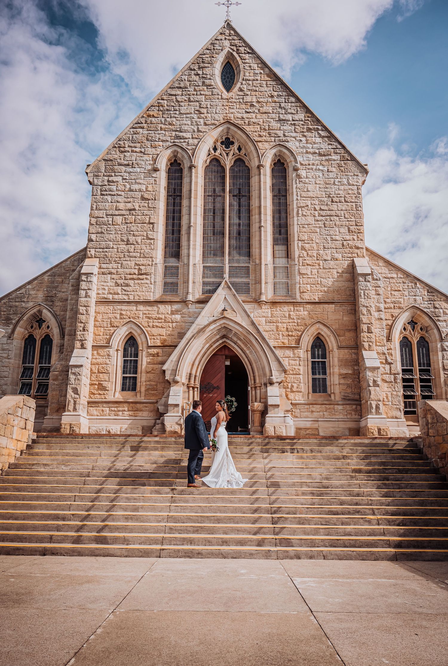 Couple standing on stone steps in front of a gothic stone church with arched windows and doorway on their wedding day.