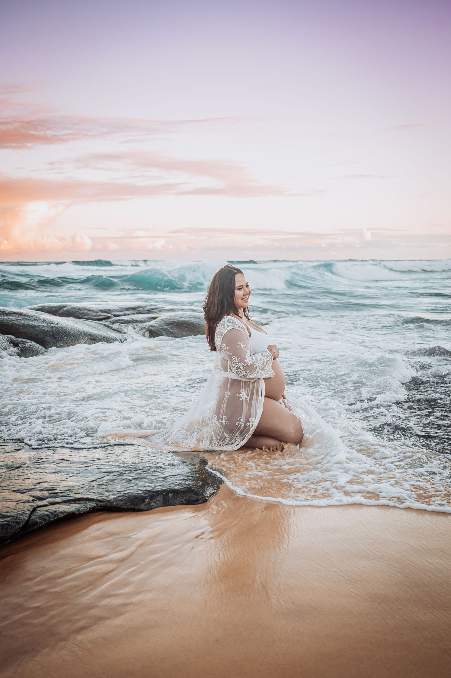 White lace dress flowing in the ocean waves during a pastel sunset on a rocky beach.