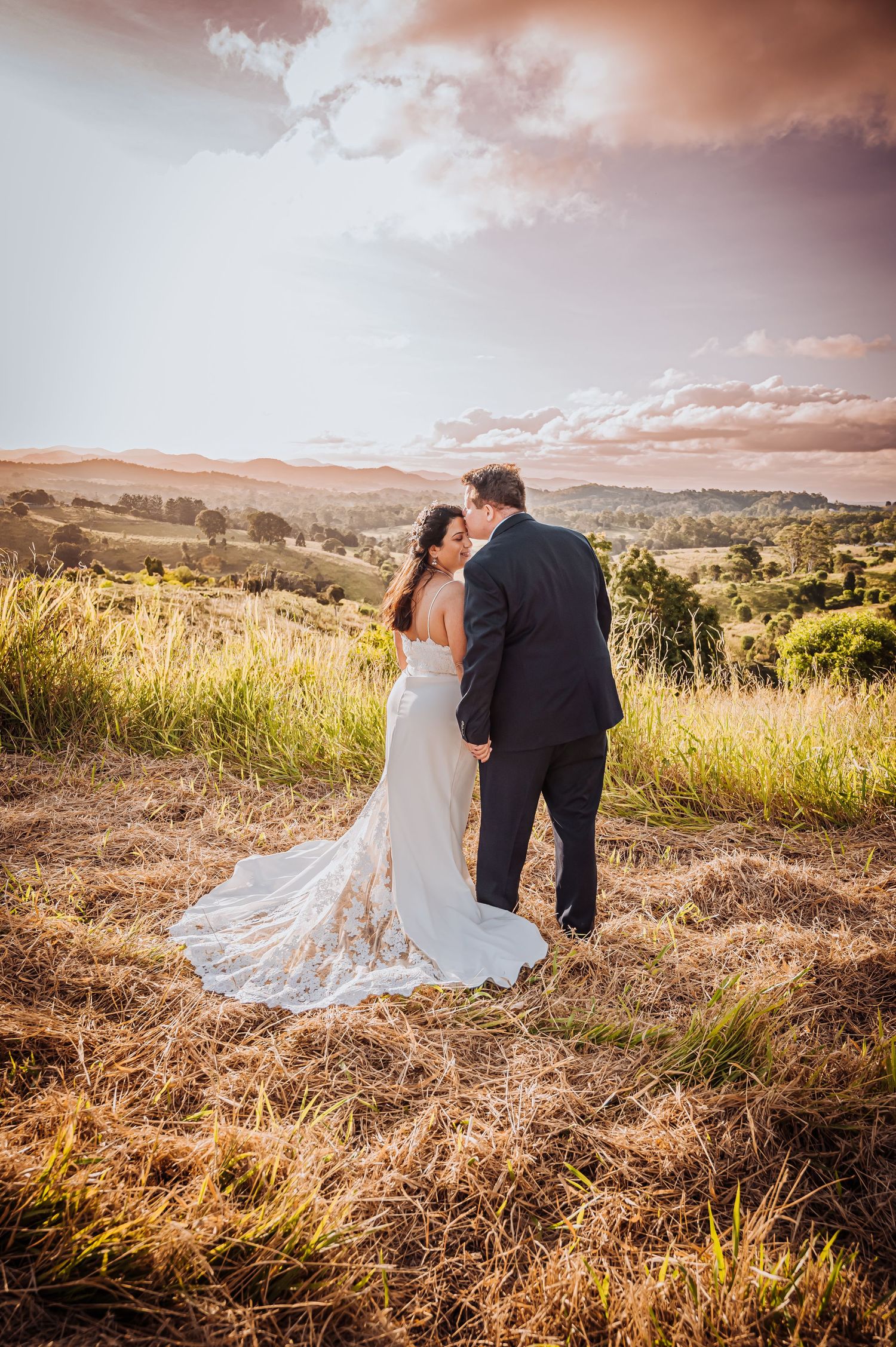 A wedding couple stands on a hilltop overlooking a scenic valley at sunset.
