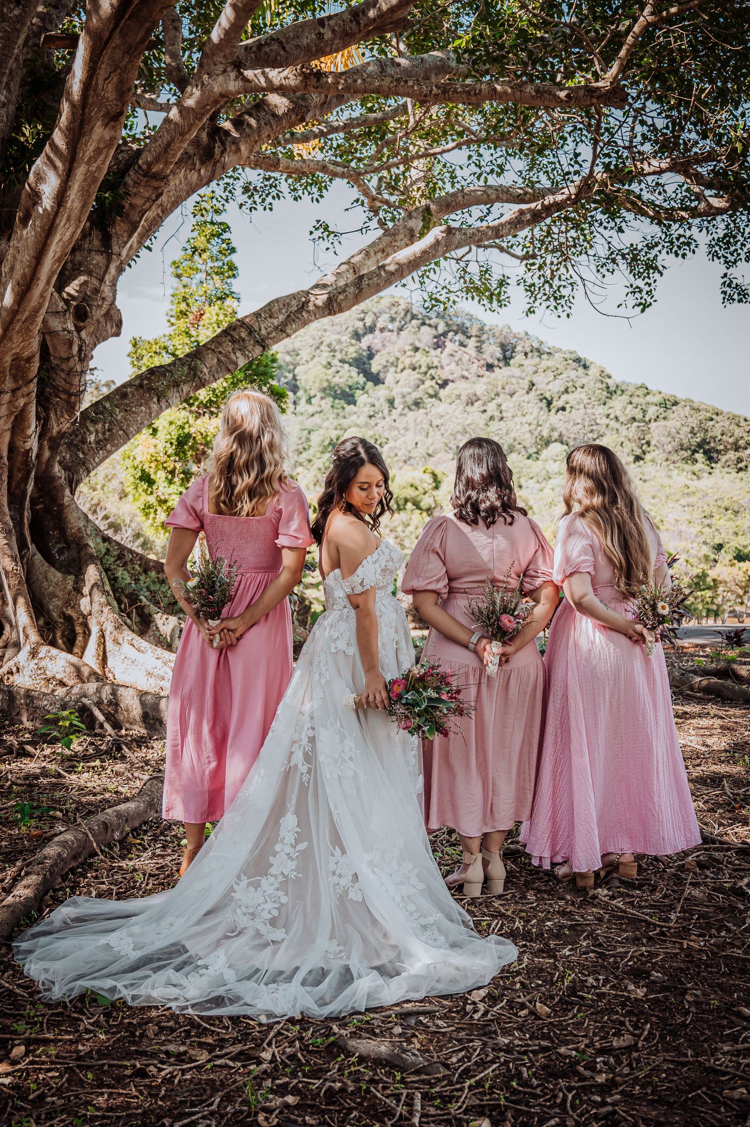 Wedding party poses under a large tree with bridesmaids in pink dresses and a bride in an elegant white gown.